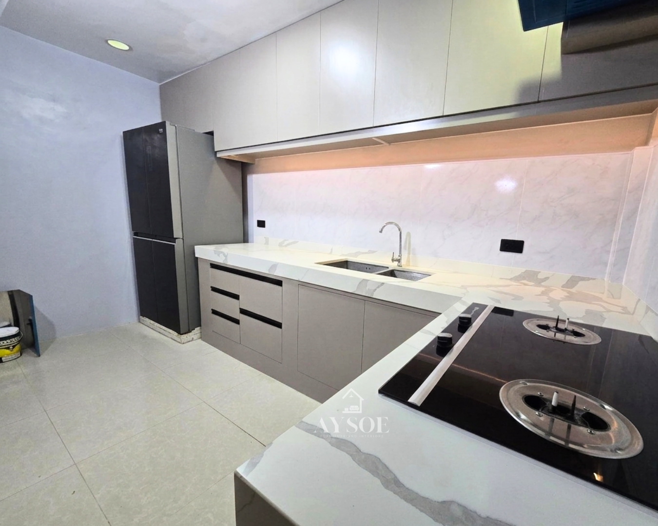 A modern L-shaped kitchen set with cream upper cabinets, taupe lower cabinets, and a marble-look countertop. Handleless, bright, and full of storage.