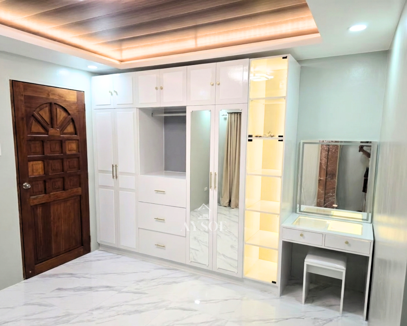 A floor-to-ceiling white shaker wardrobe with gold hardware, mirror doors, and drawers. A lit glass display tower and built-in vanity complete the wall.
