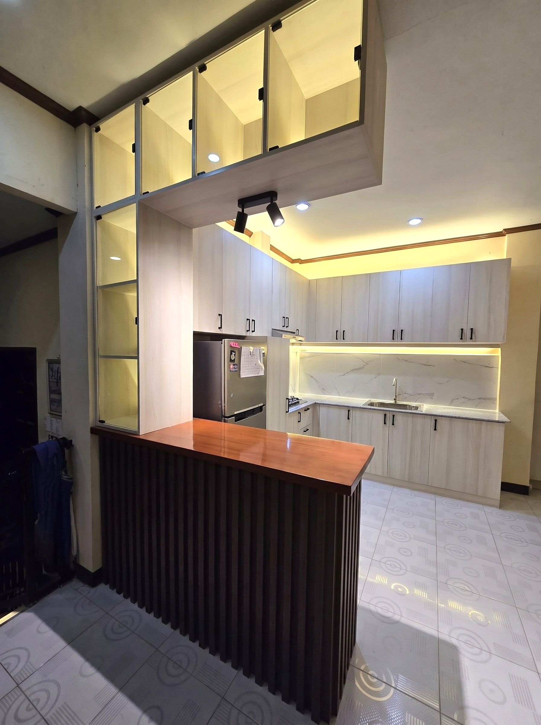 A light oak L-shaped kitchen with a marble-look backsplash, wood breakfast bar, and lit glass display cabinets.