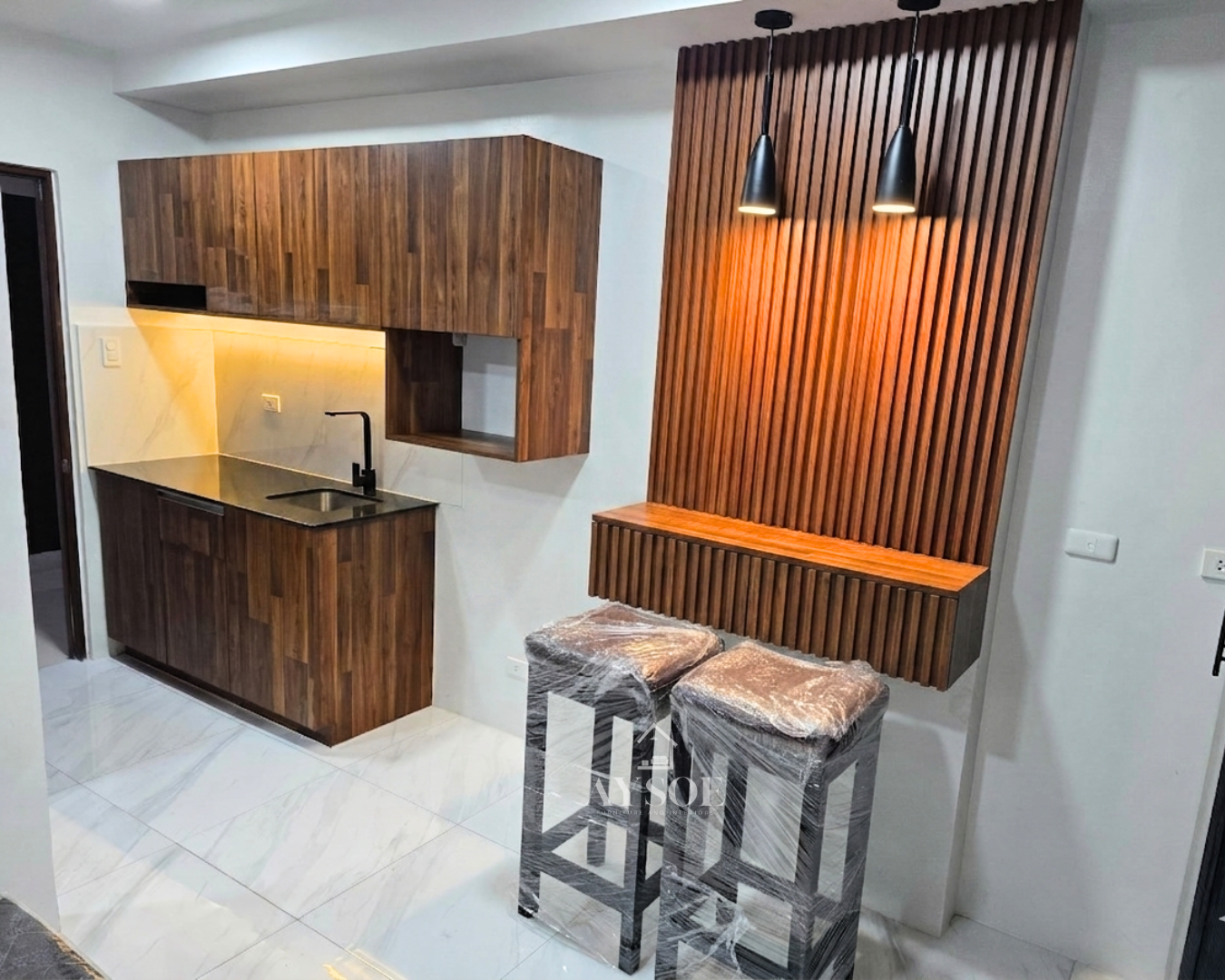 A compact walnut-finish kitchenette with upper and lower cabinets, plus a floating bar counter with a fluted wood wall panel and pendant lights. Made for condos and studio units.