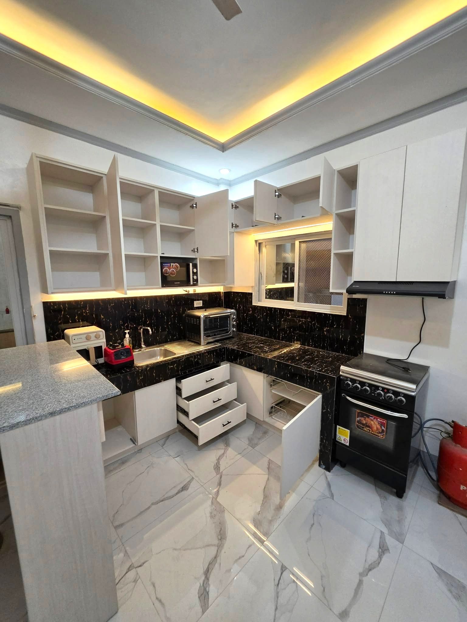 A cream wood-grain L-shaped kitchen with a black and gold backsplash, open shelves, and a granite-top breakfast bar.