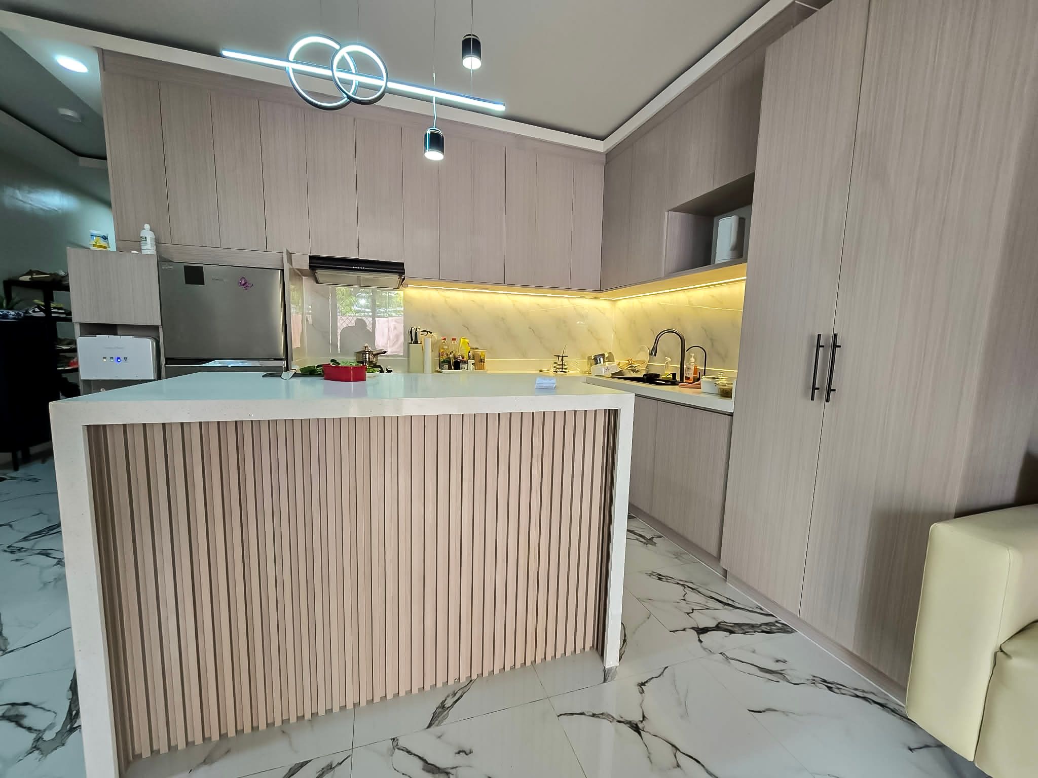 An L-shaped kitchen in soft taupe wood-grain cabinets, with a marble-look backsplash, warm LED lighting, and a white waterfall island with a wood-slat front. Tall storage and cabinets up to the ceiling make the most of the space.