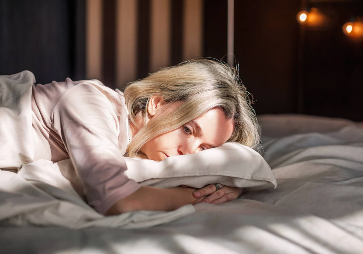 Mujer rubia tumbada boca abajo abraza una almohada con mirada pensativa