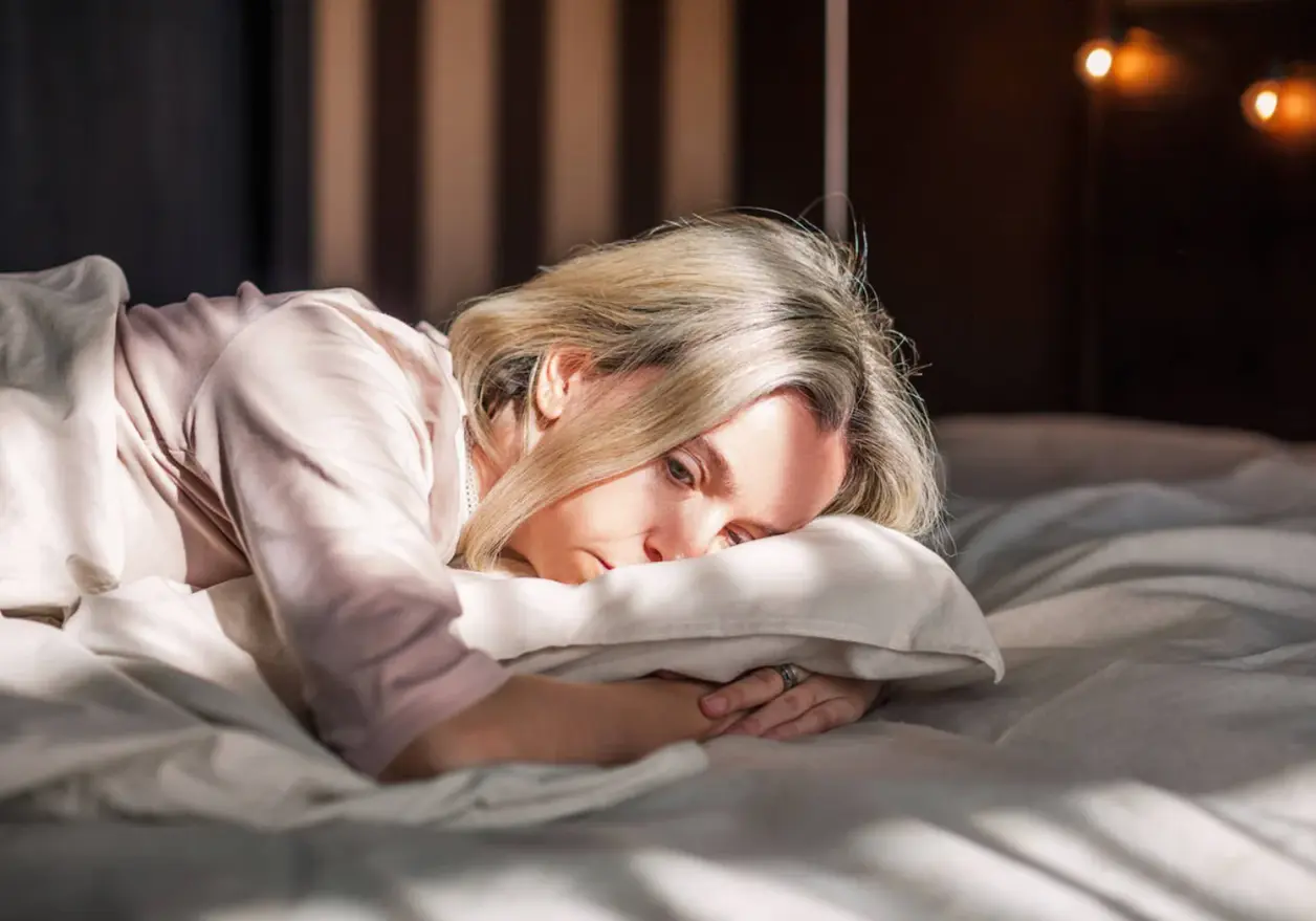 Mujer rubia tumbada boca abajo abraza una almohada con mirada pensativa