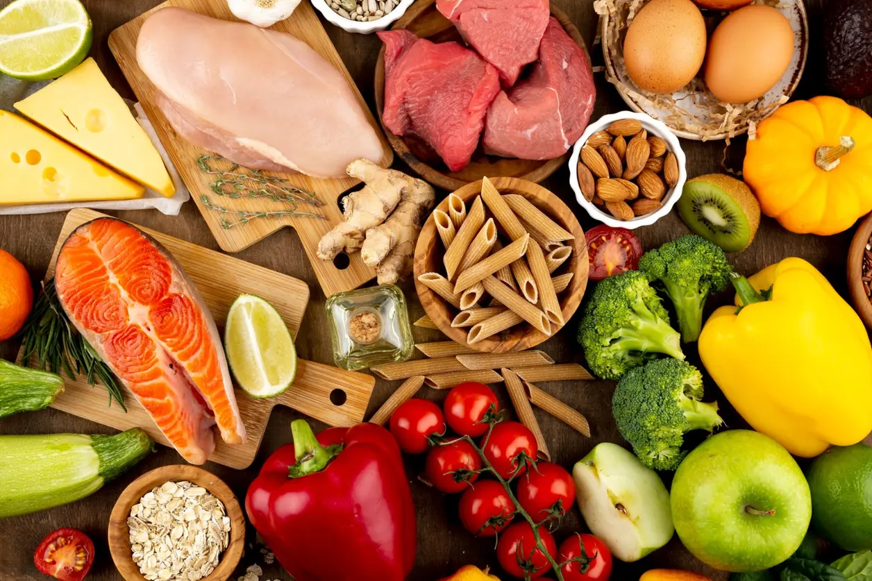 Mesa llena de alimentos frescos: salmón, carne, huevos, verduras, frutas, frutos secos y pasta integral