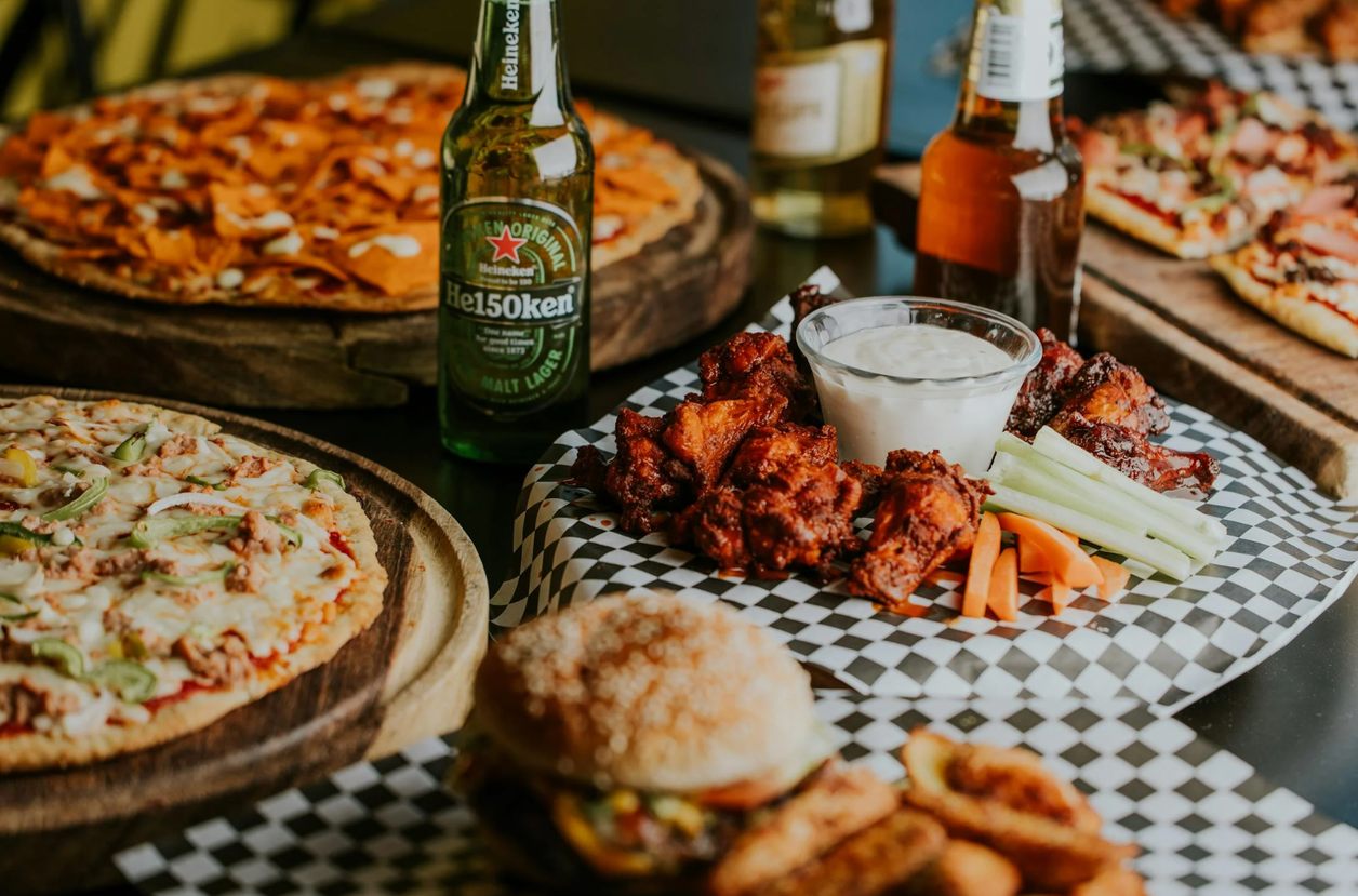 Mesa con pizzas, alitas de pollo, hamburguesa y botellas de cerveza