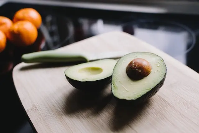 Aguacate cortado por la mitad sobre una tabla, con mandarinas al fondo