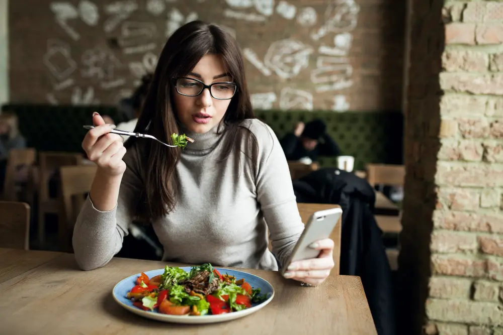 Mujer con gafas come ensalada mientras mira el móvil en una cafetería