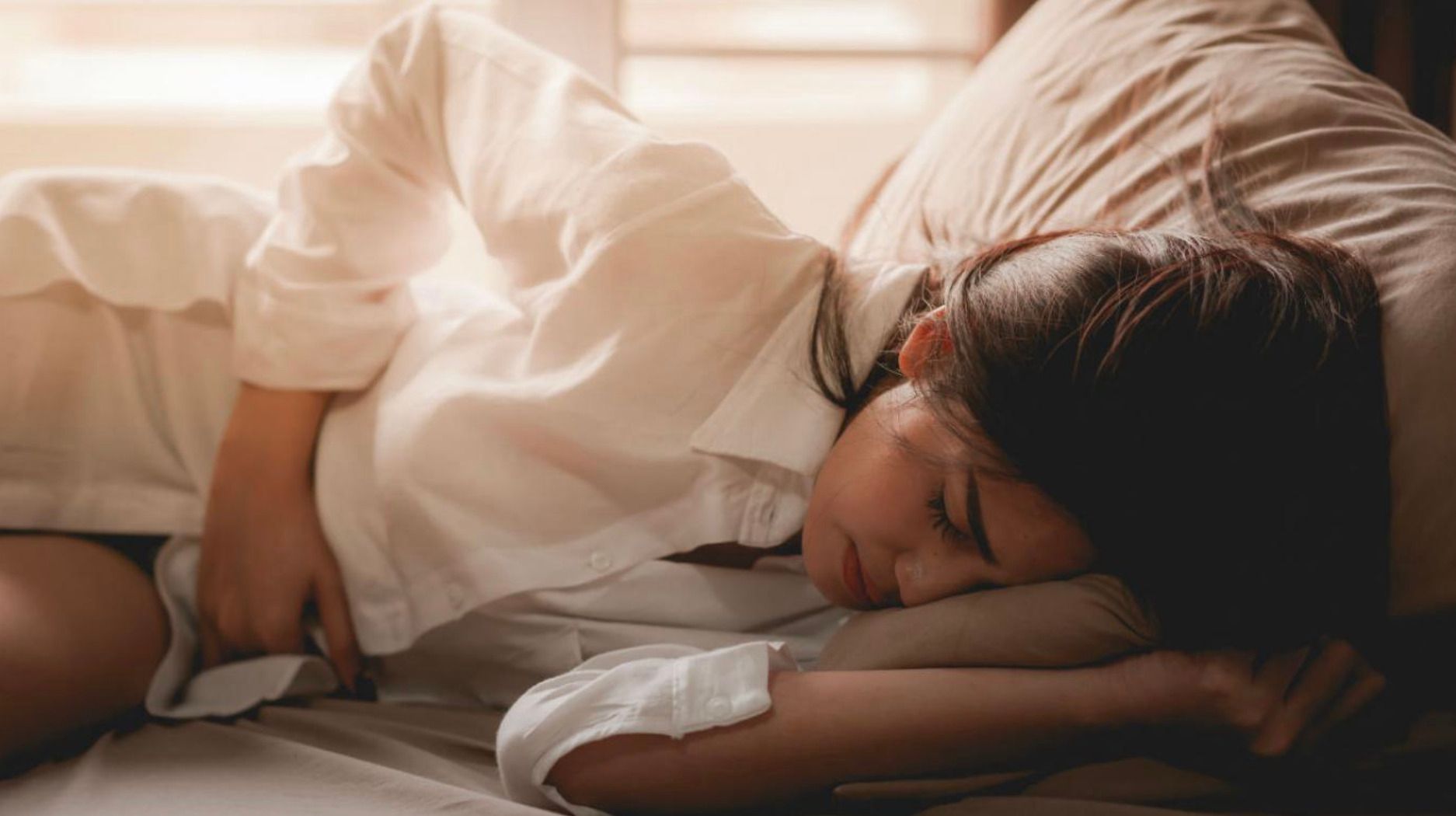 Mujer tumbada de lado en la cama se encoge de dolor con las manos en el vientre
