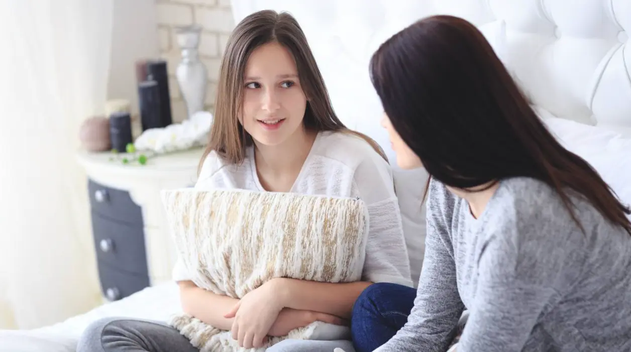 Adolescente abraza un cojín mientras habla con su madre sentadas en la cama