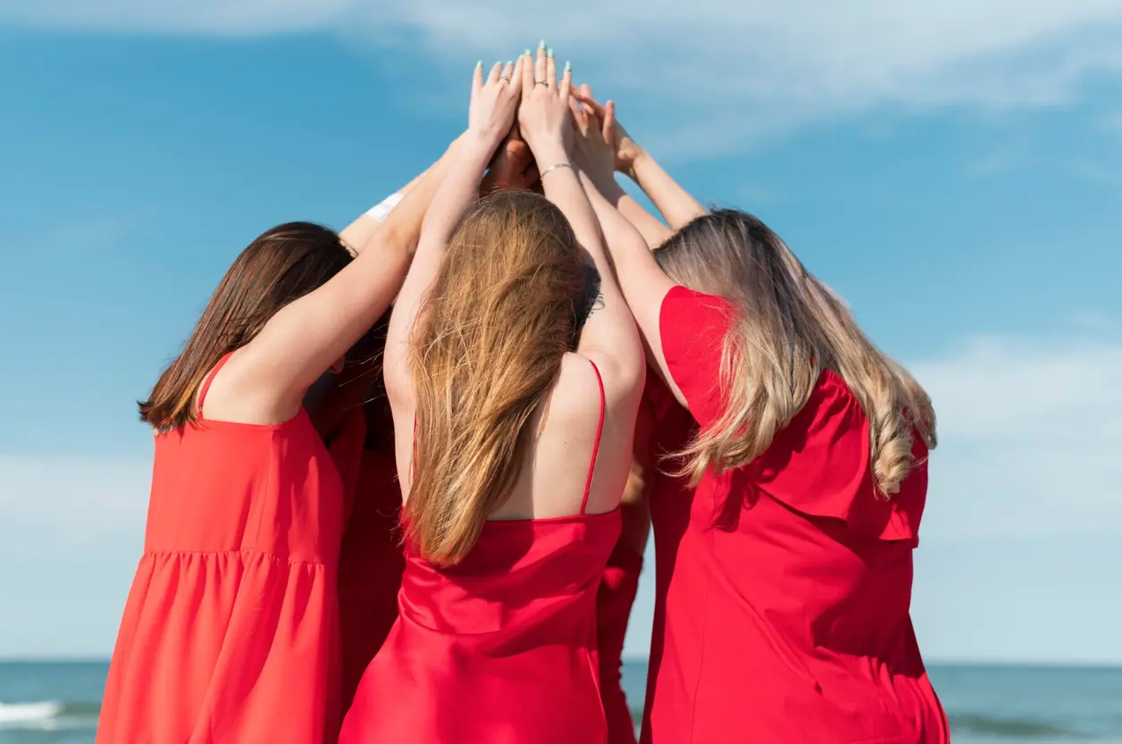 Tres amigas con vestidos rojos juntan las manos en alto frente al mar