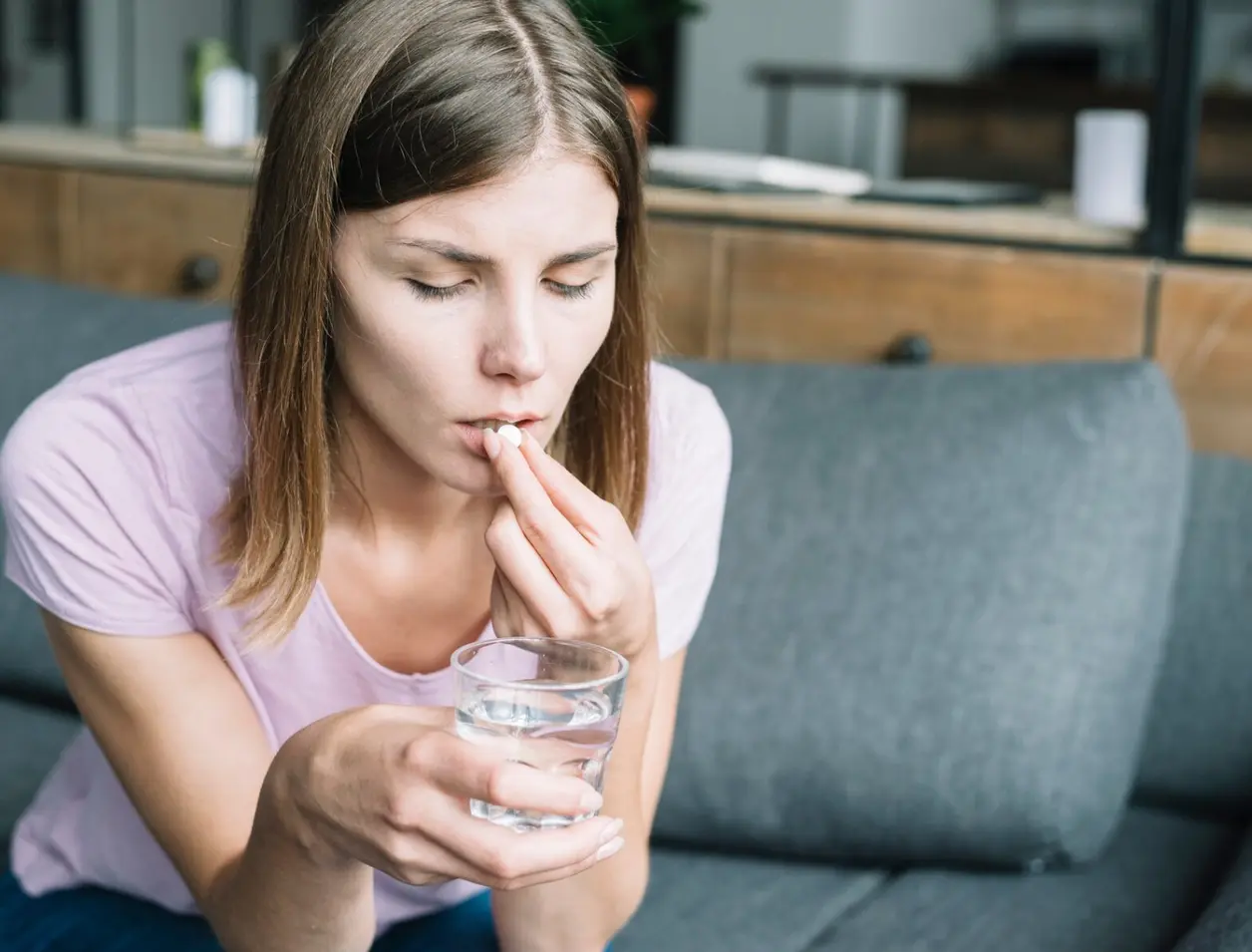 Mujer sentada en un sofá toma una pastilla con un vaso de agua