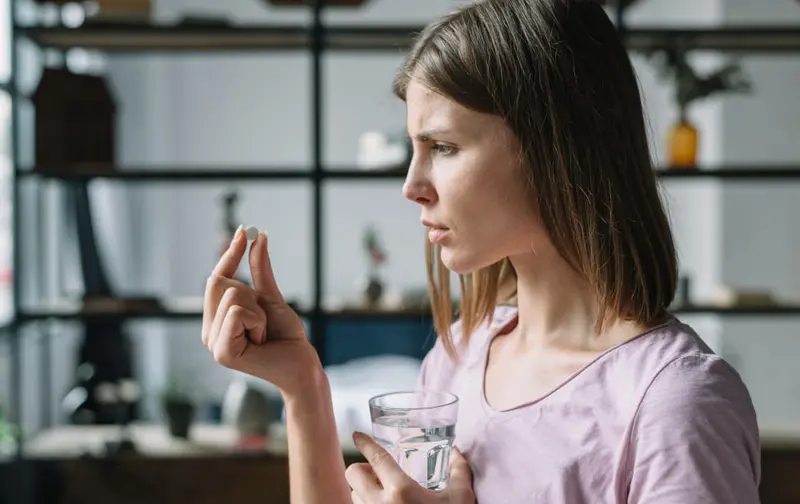 Mujer de perfil observa una pastilla mientras sostiene un vaso de agua