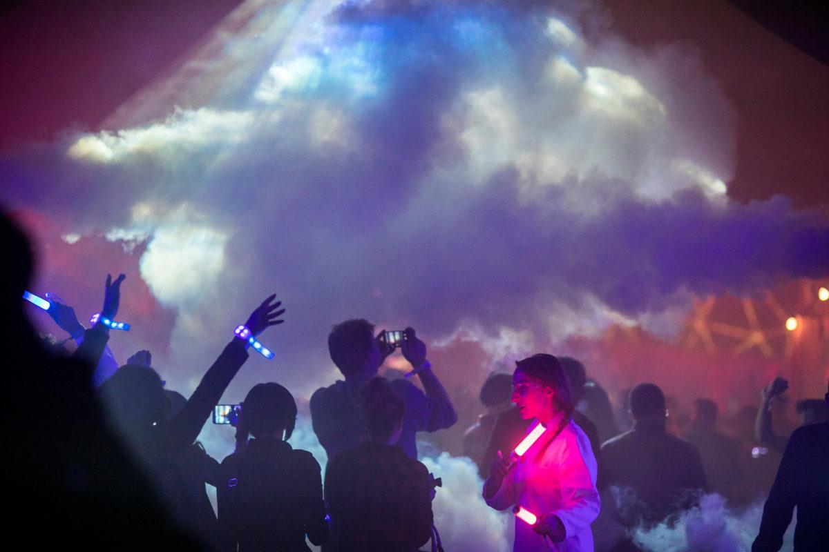 Fog bursts onto the dancefloor as partygoers with glowing wristbands photograph it.