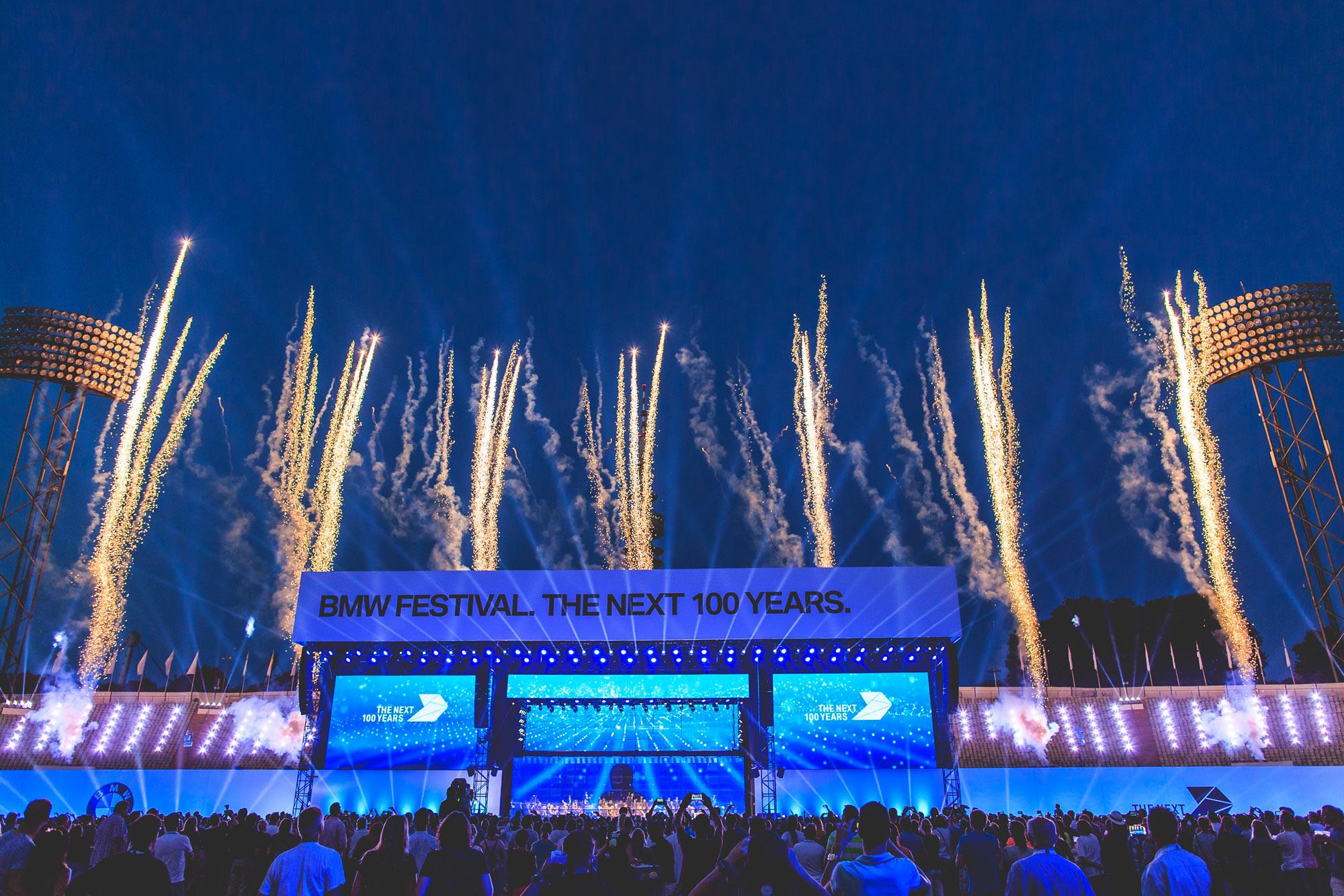 Fireworks burst upwards into a blue dusky sky above a stage that reads "BMW FESTIVAL. THE NEXT 100 YEARS." and a large crowd.