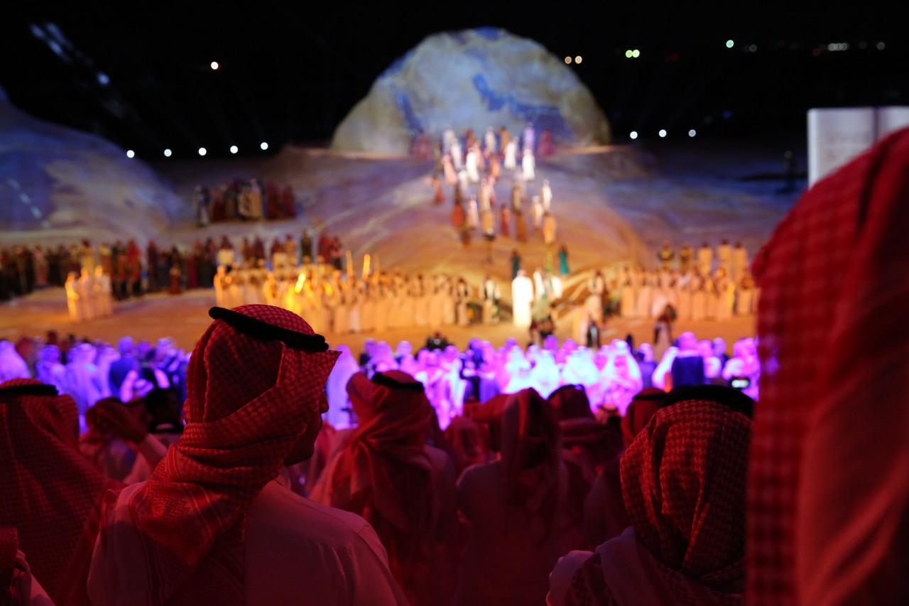 The large stage is filled with performers in neat formations as a large crowd looks on.