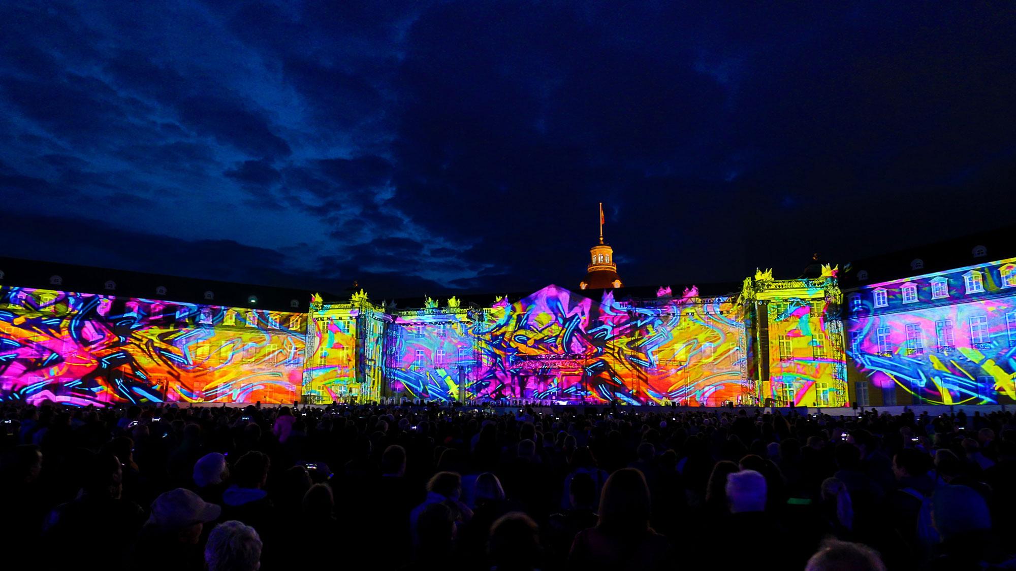 Projection mapping of bright neon graffiti patterns on a building. A sea of people looks on.