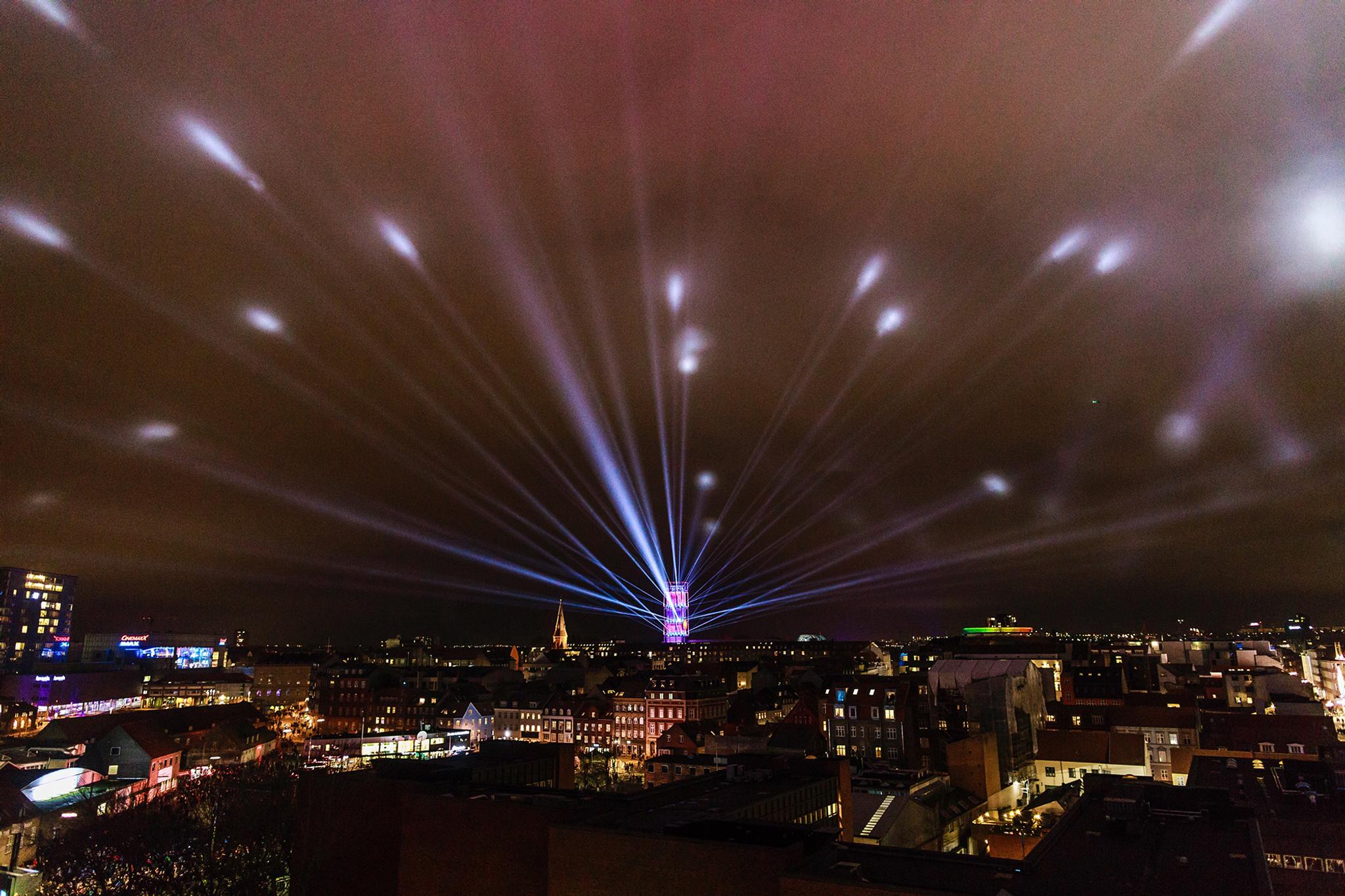 A broad view of Aarhus from above at night. in the center, a tower of light emits long reaching beams into the night sky, stretching all across the city.