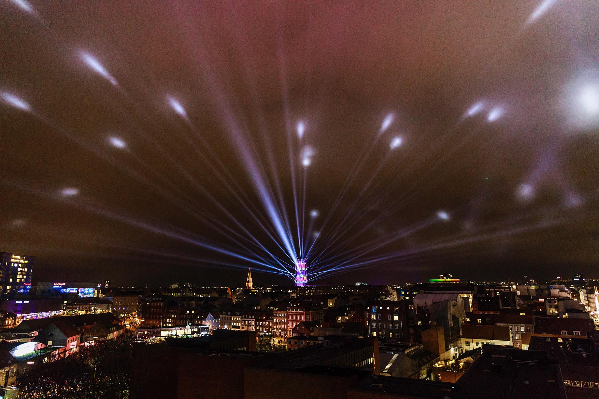 A broad view of Aarhus from above at night. in the center, a tower of light emits long reaching beams into the night sky, stretching all across the city.