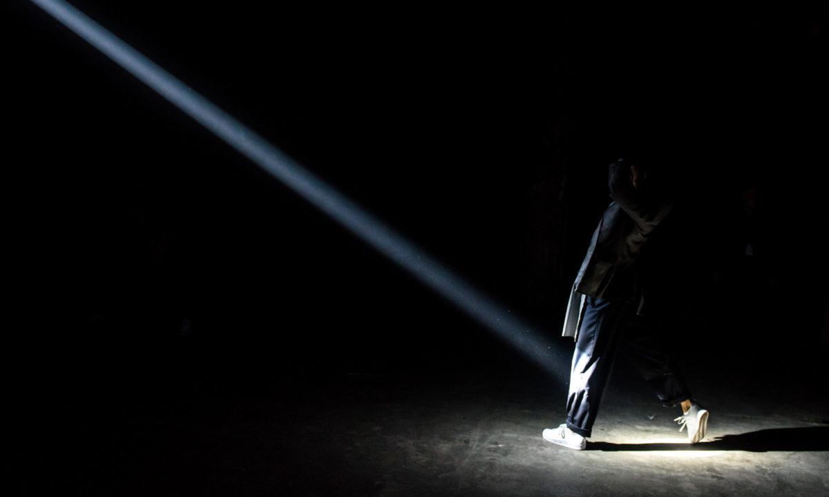 A figure walks into a single beam of light in an otherwise completely black frame. They are dressed in black and wear white sneakers.