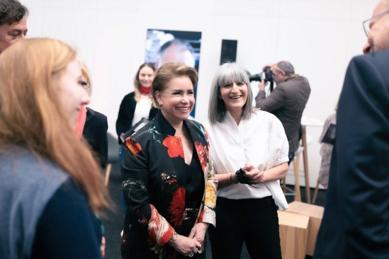 Nancy Braun and a colleague stand in the center of frame and socialize in a group in a gallery space.