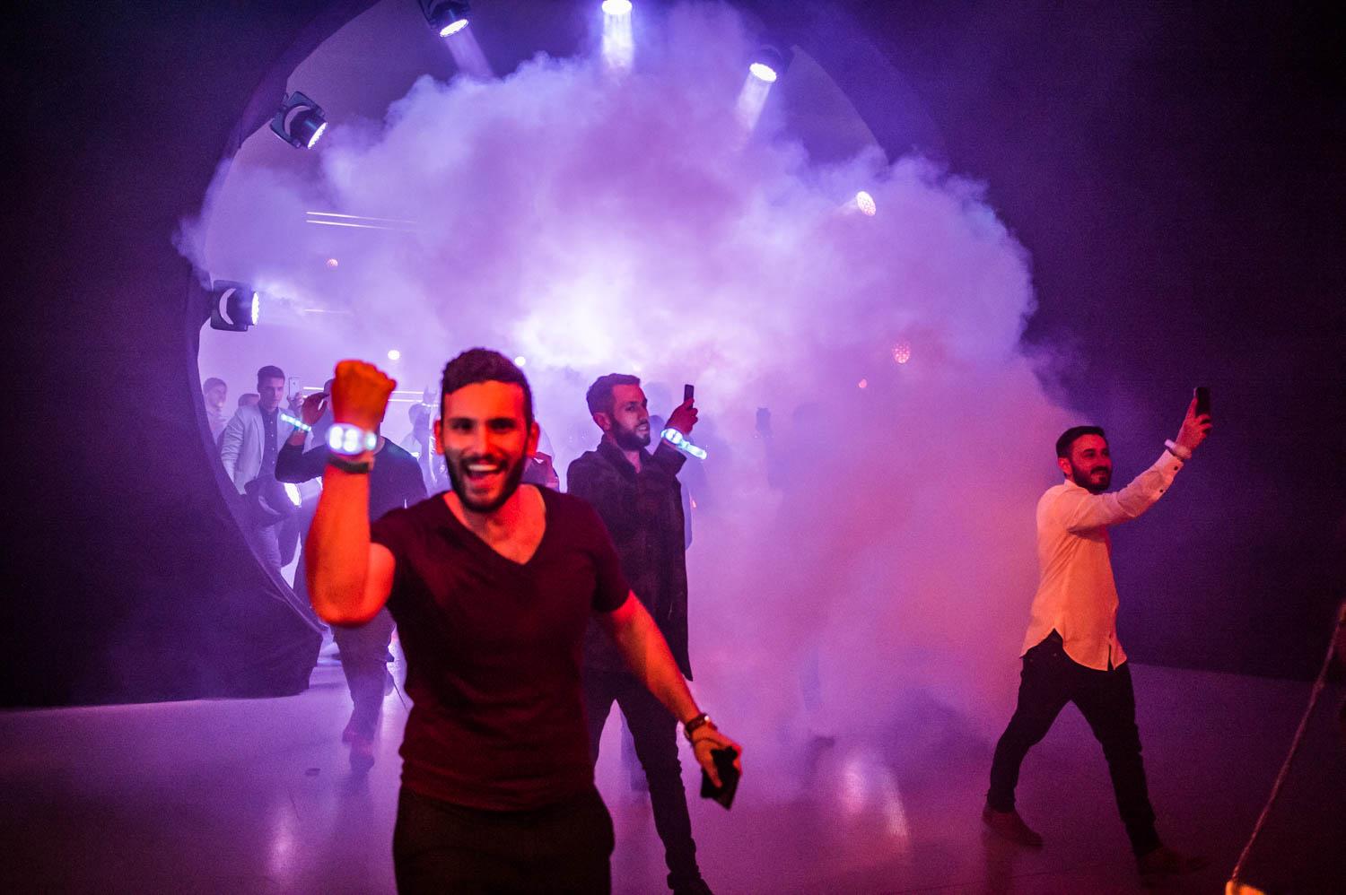 A group of party-goers emerges through a cloud of fog. A man faces the camera showing off his glowing wristband and smiles with an open mouth. Other party goers walk while filming with their phones.