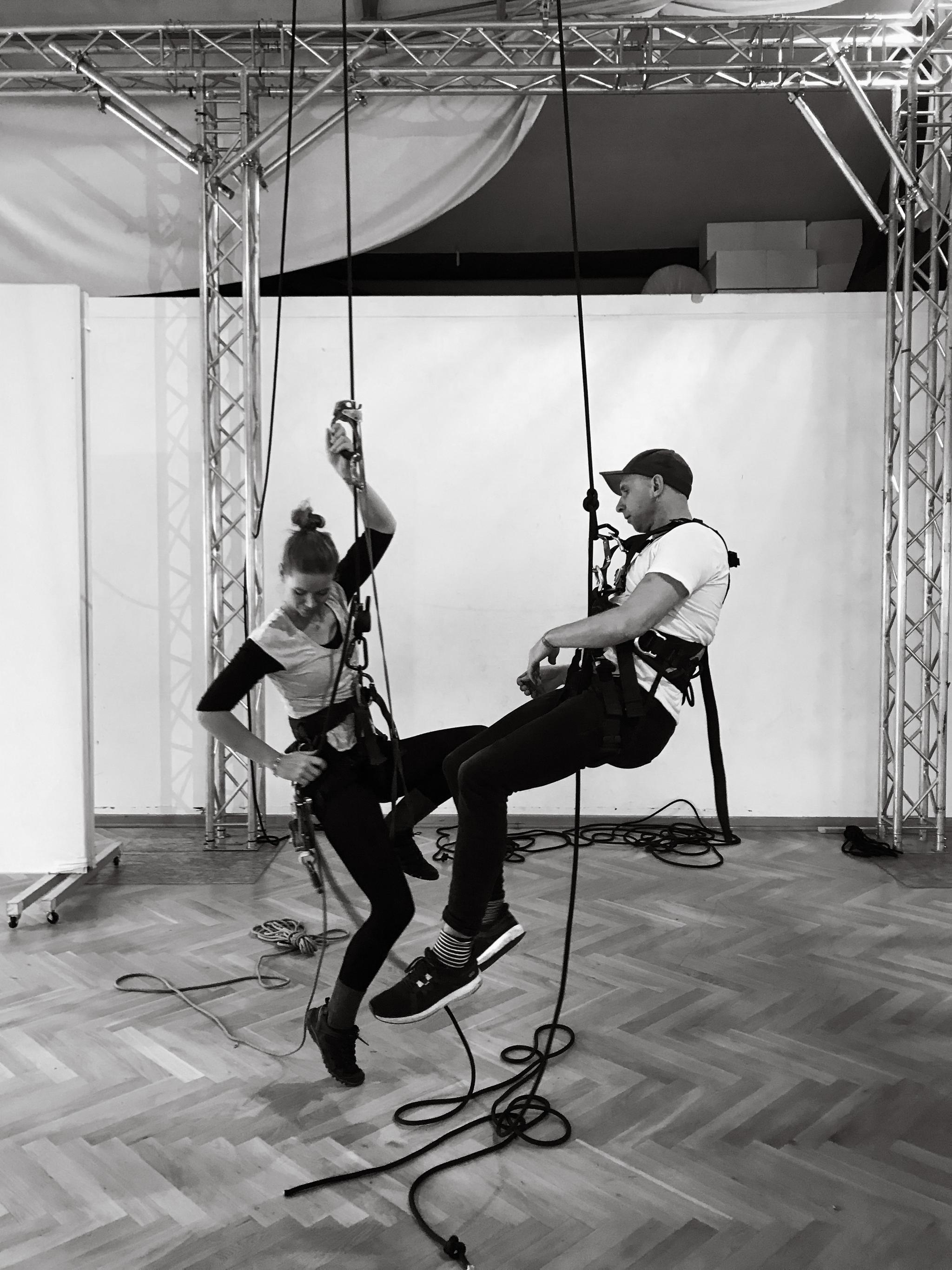 Two people hang close to a wood parquet floor from black ropes tied to metal trusses. They are secured with carabeeners and harnesses. The photo is black and white.