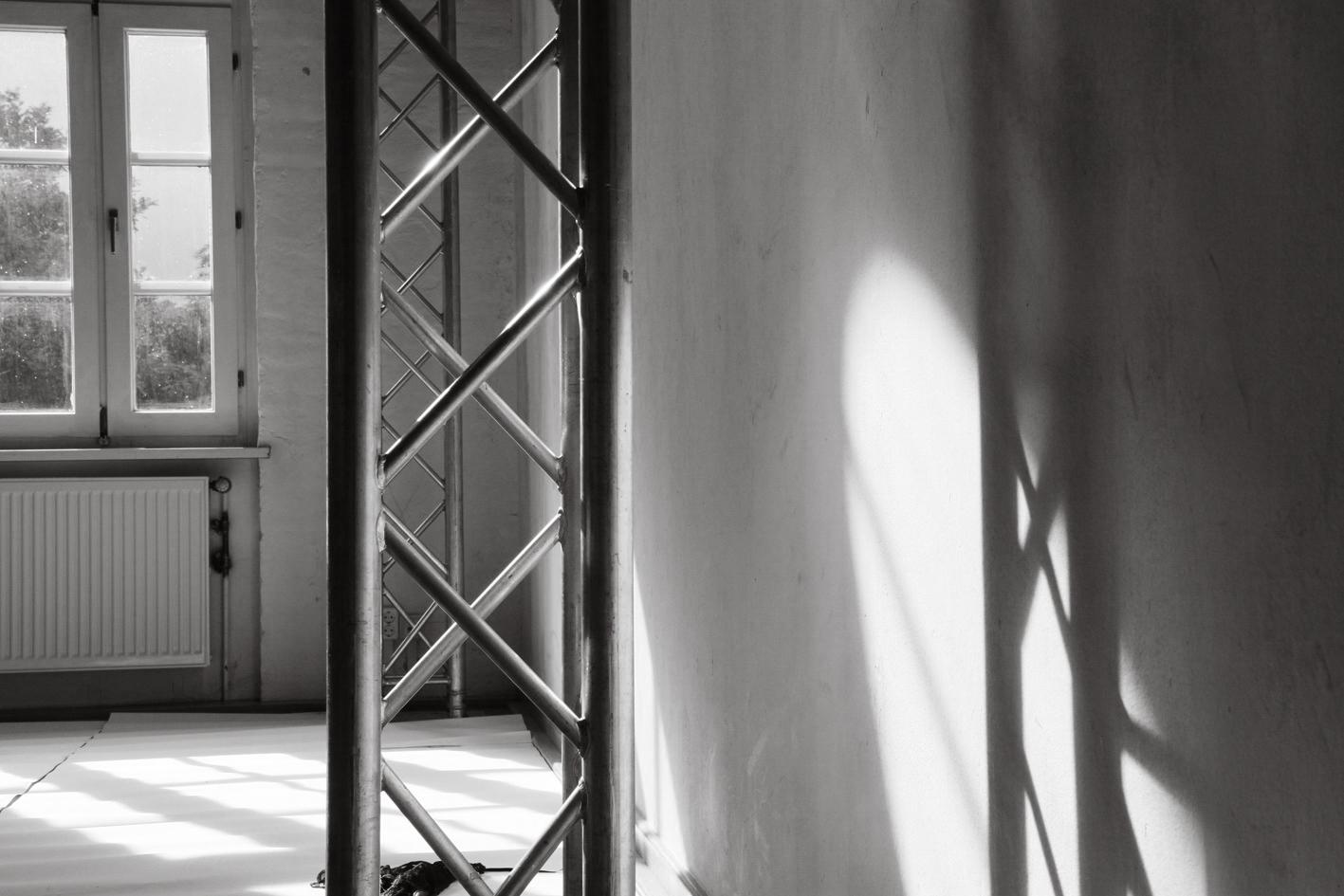 Afternoon sun shines through an altbau window and gleams off of metal trusses in the front of the frame, casting soft shadows onto the wall. The image is black and white.