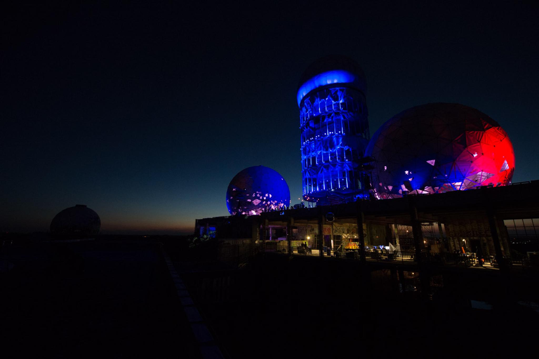 The last traces of the sun are barely hanging onto the horizon. The sky is a deep, dark blue. The domes, and a tower on Teufelsberg are lit in blue and red.