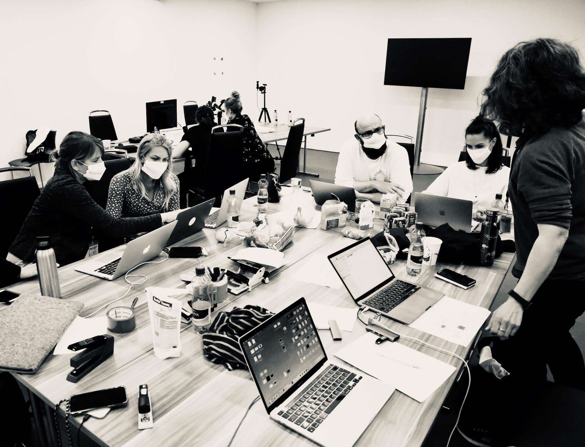 A black and white photo of 6 people wearing masks on laptops at a wooden table.