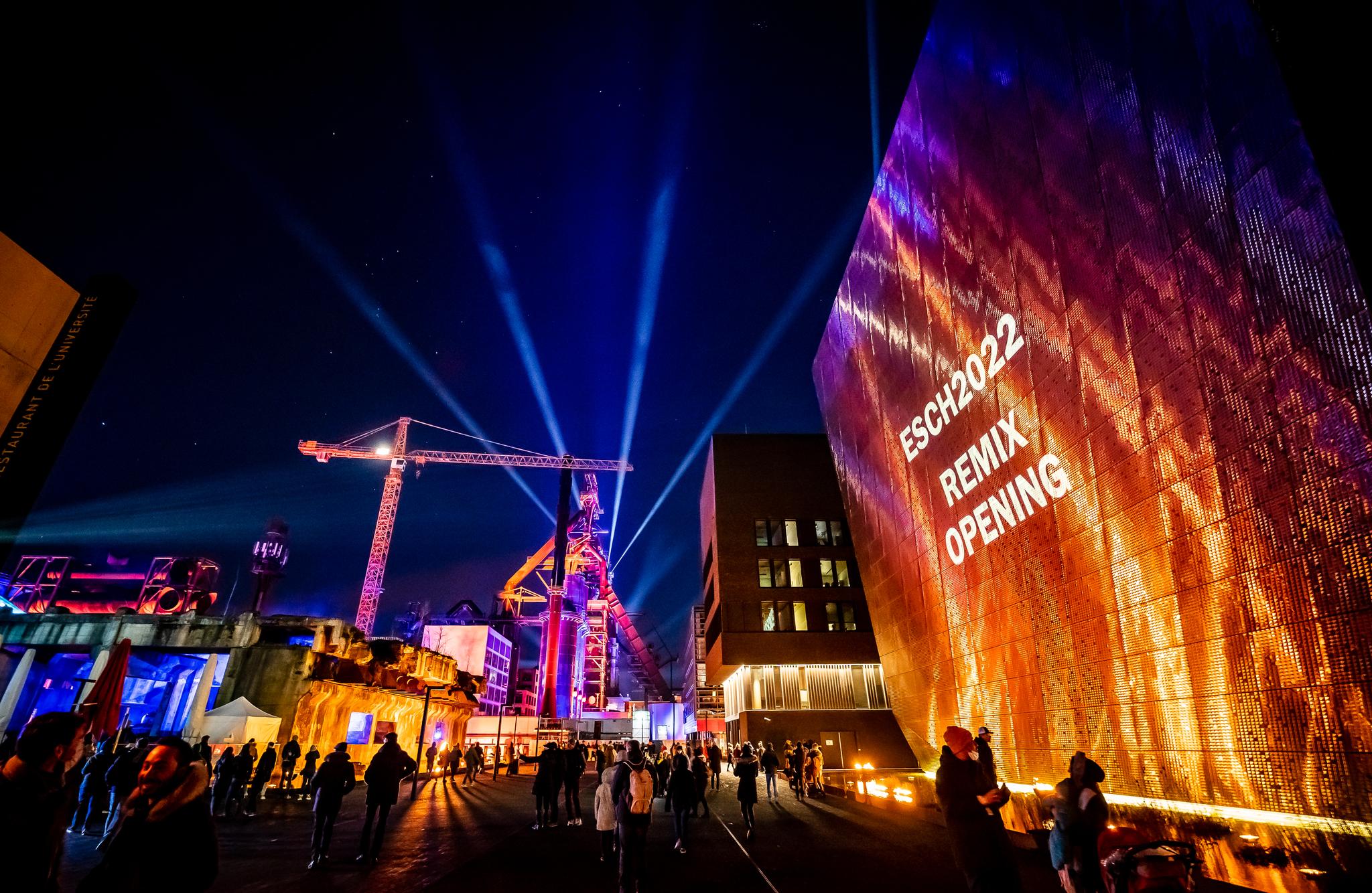 Outdoor scene at night. "ESCH2022 REMIX OPENING" is projected on a large stylized wall. A crane and spotlights are in the background and participants mill about.
