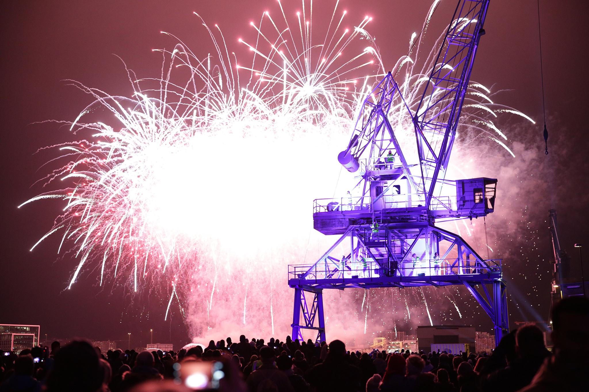 Big bursts of fireworks in front of a purple-lit crane. A crowd watches on.
