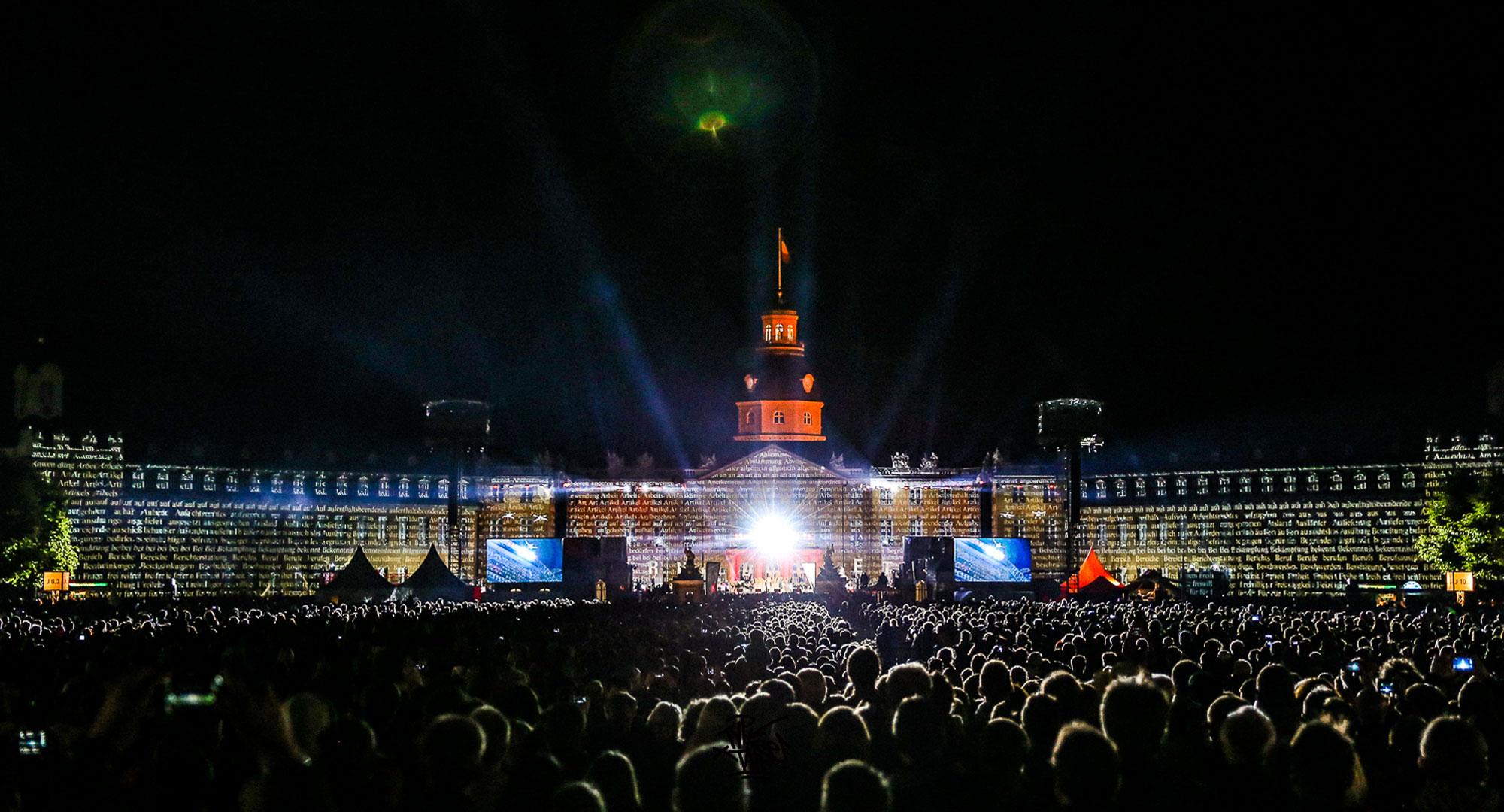 A sea of people watch video mapping on a building. There is a performance on stage.