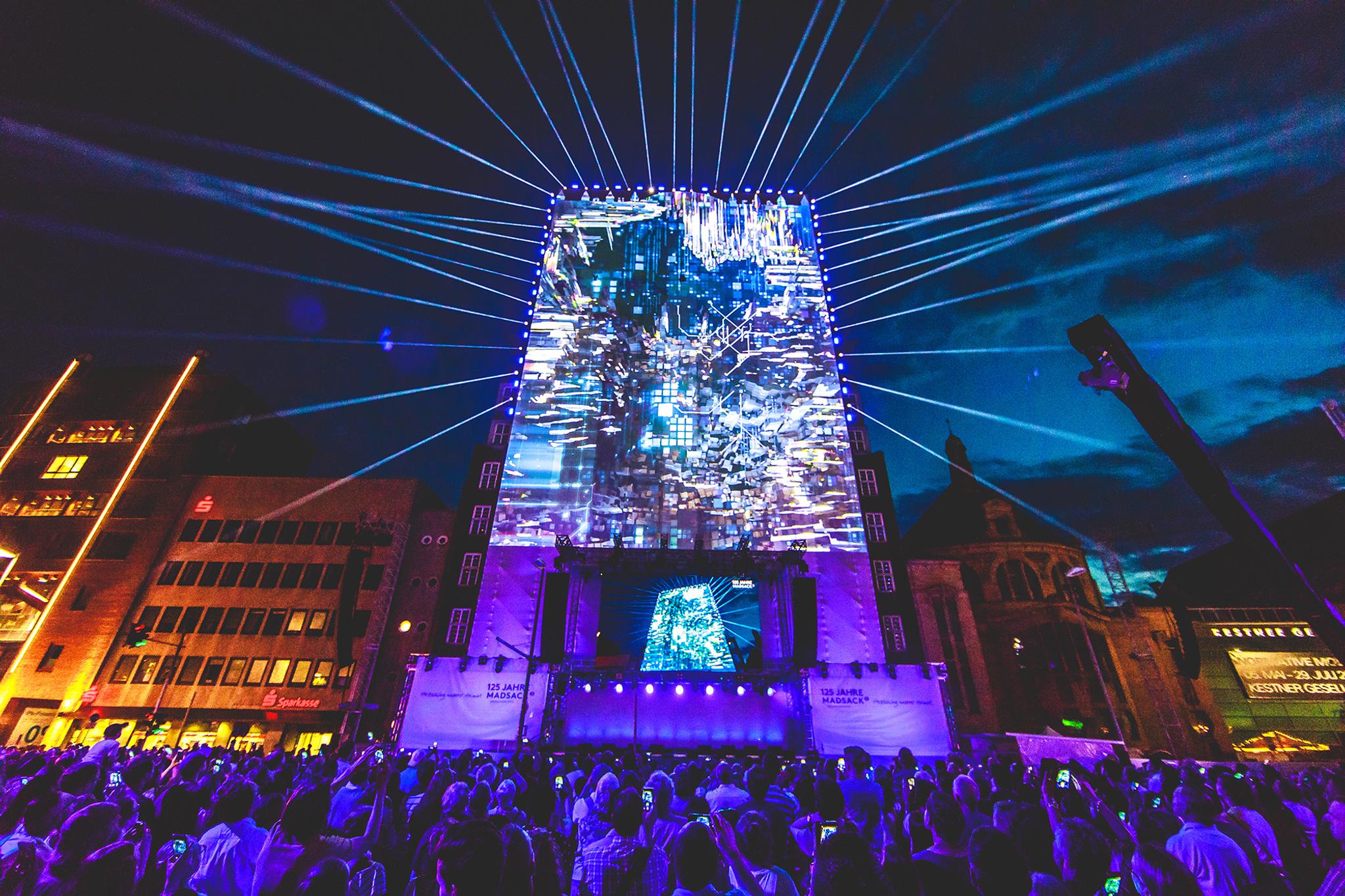 Video mapping of glitchy looking blue cubes on a large vertical stage. A large crowd watches outdoors.
