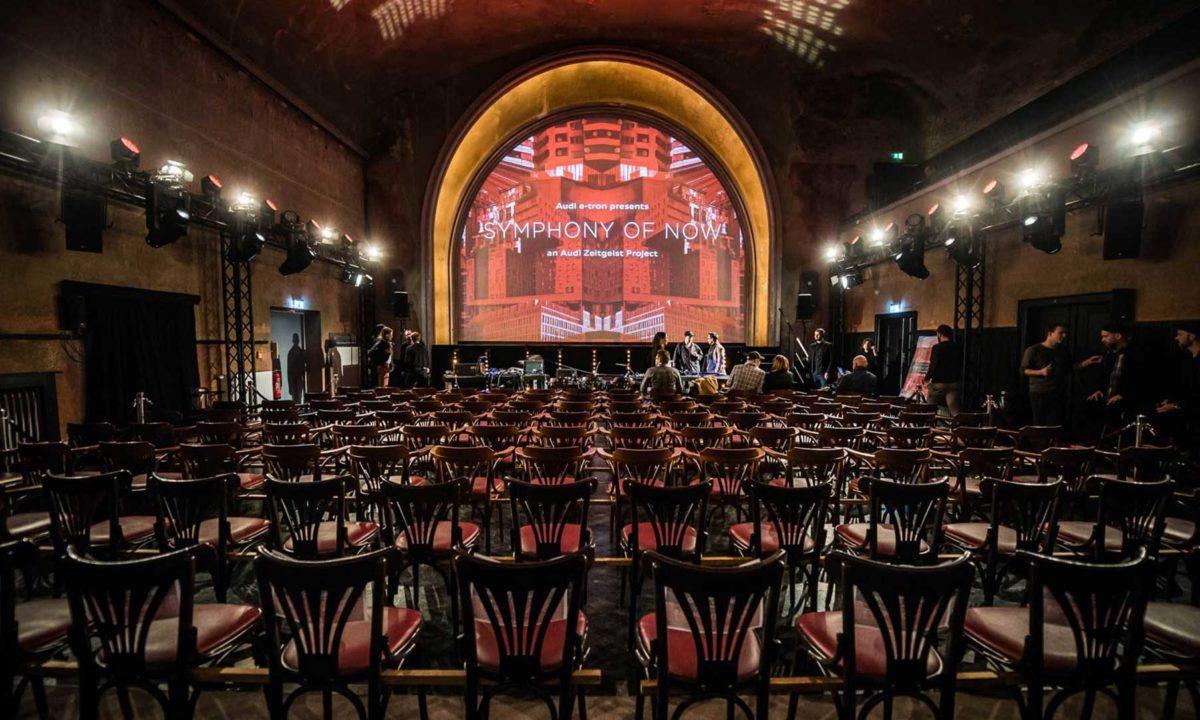 Traditional looking wooden chairs with red padding face towards an arched screen where "symphony of now" is projected.