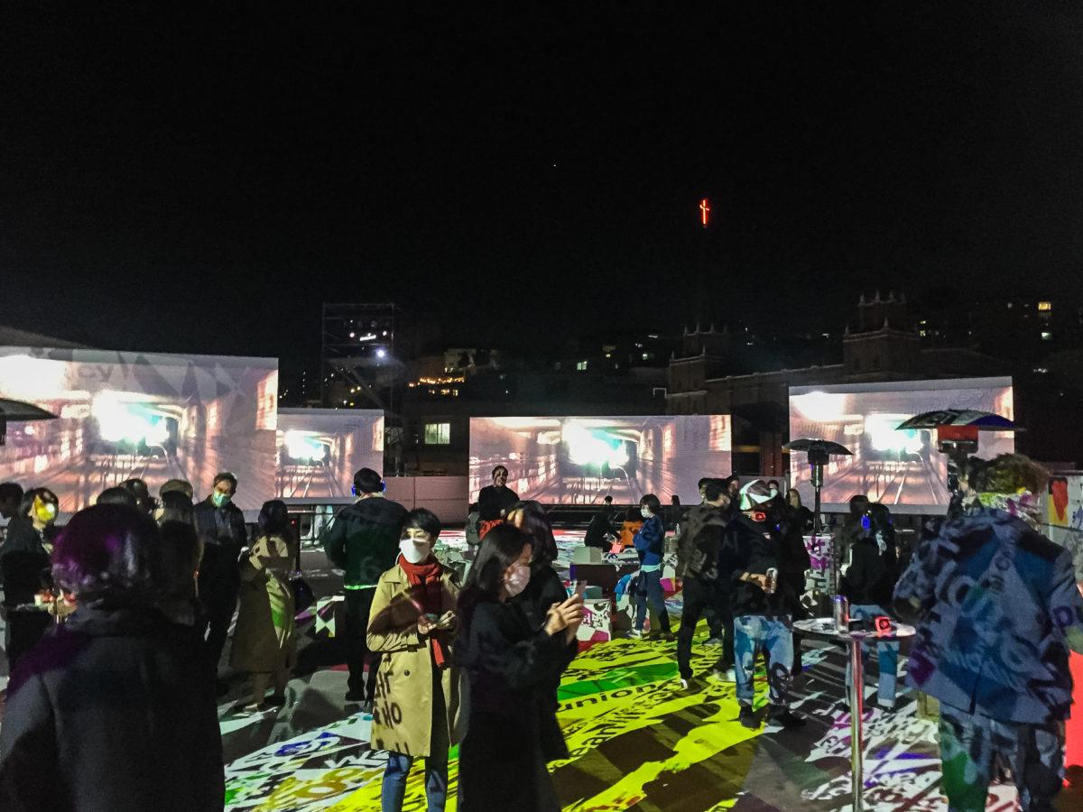 A socially distanced crowd socializes outdoors for a Symphony of Now screening. There are patterns projected on the ground.