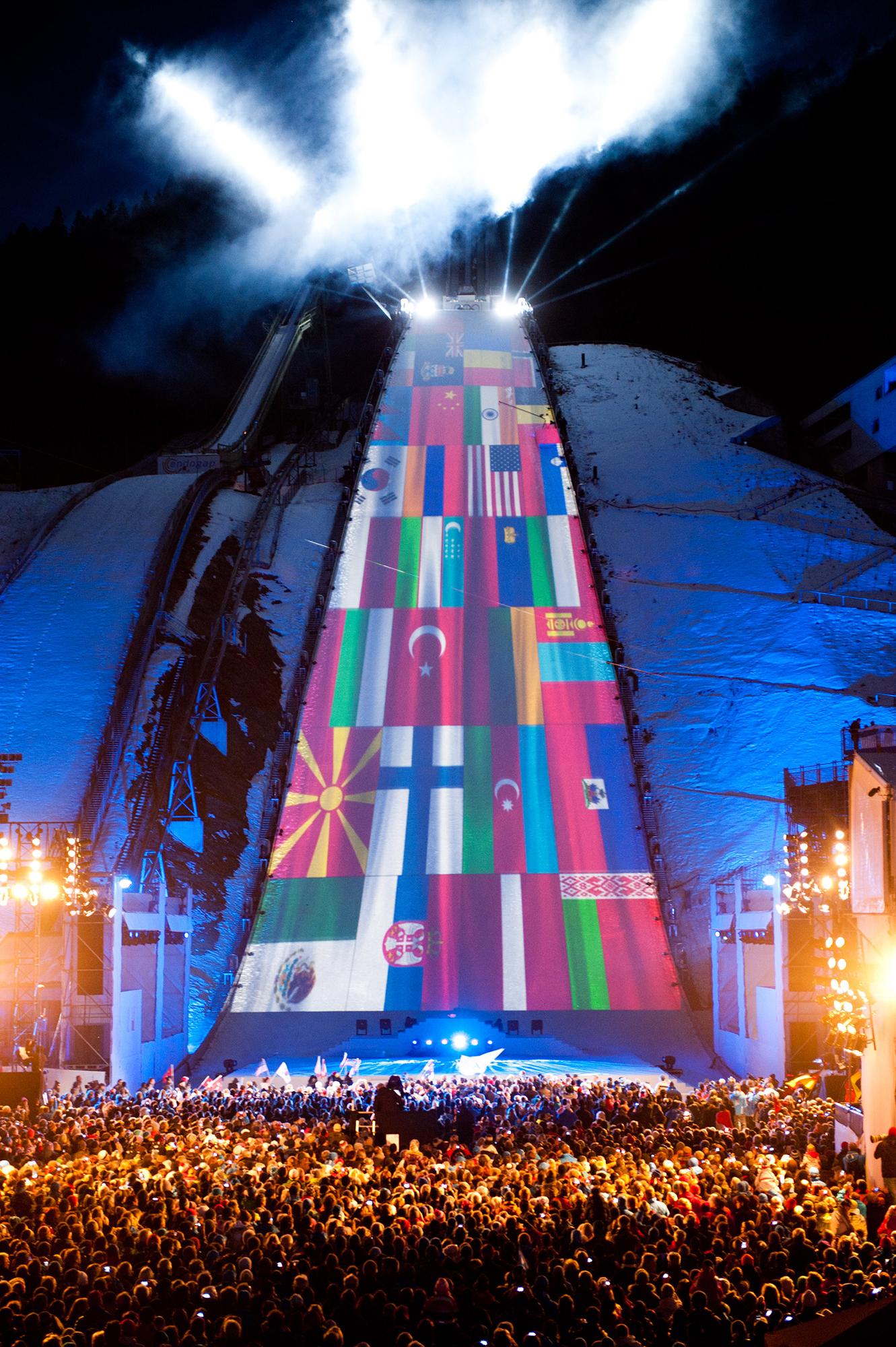 Many flags are projection mapped onto the ski slope as a sea of people looks on.