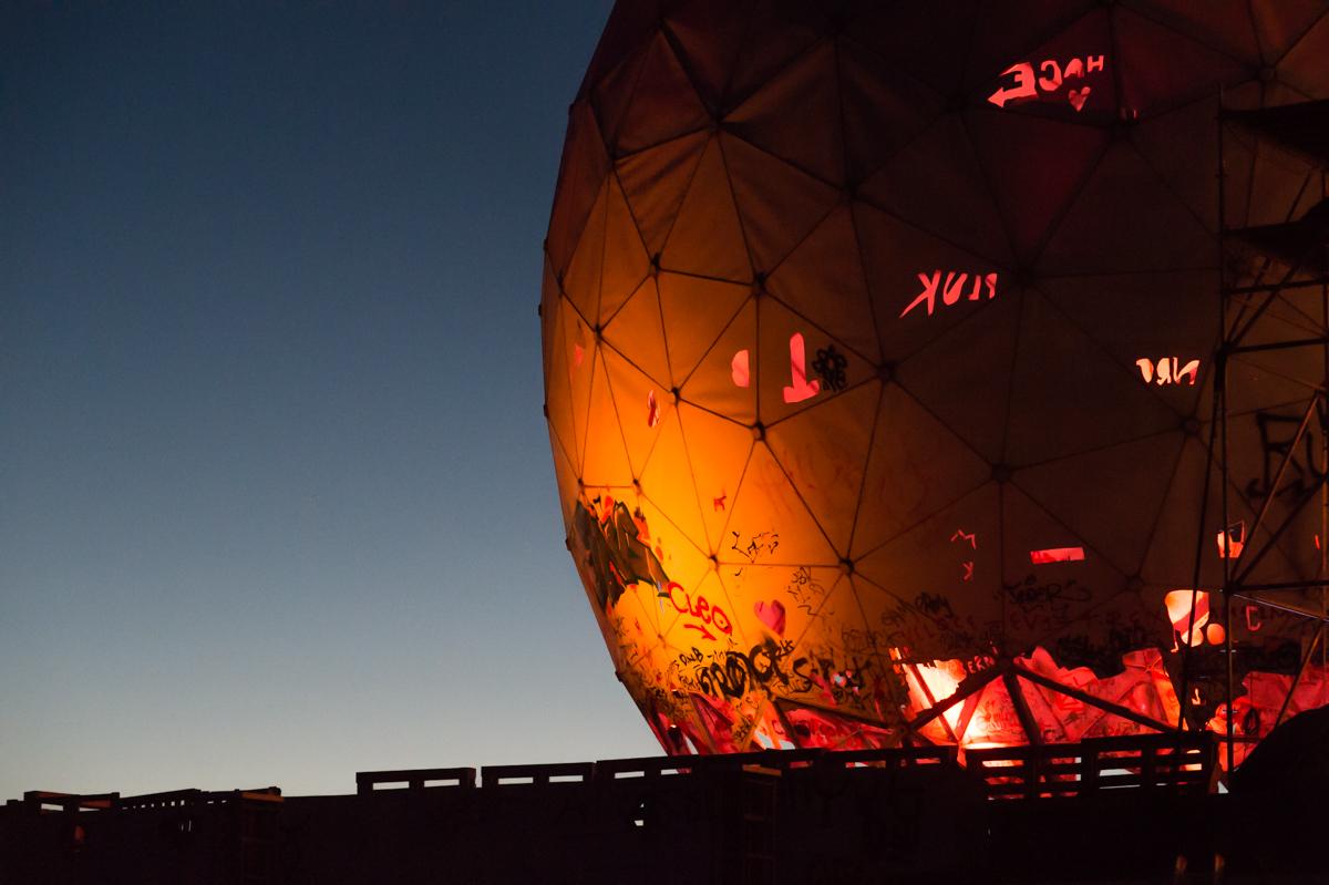 The main colors in this image are blue and a rusty orange. The left half is a clear dusky sky transitioning into blue-hour. The right half is a warmly lit dome of Teufelsberg.