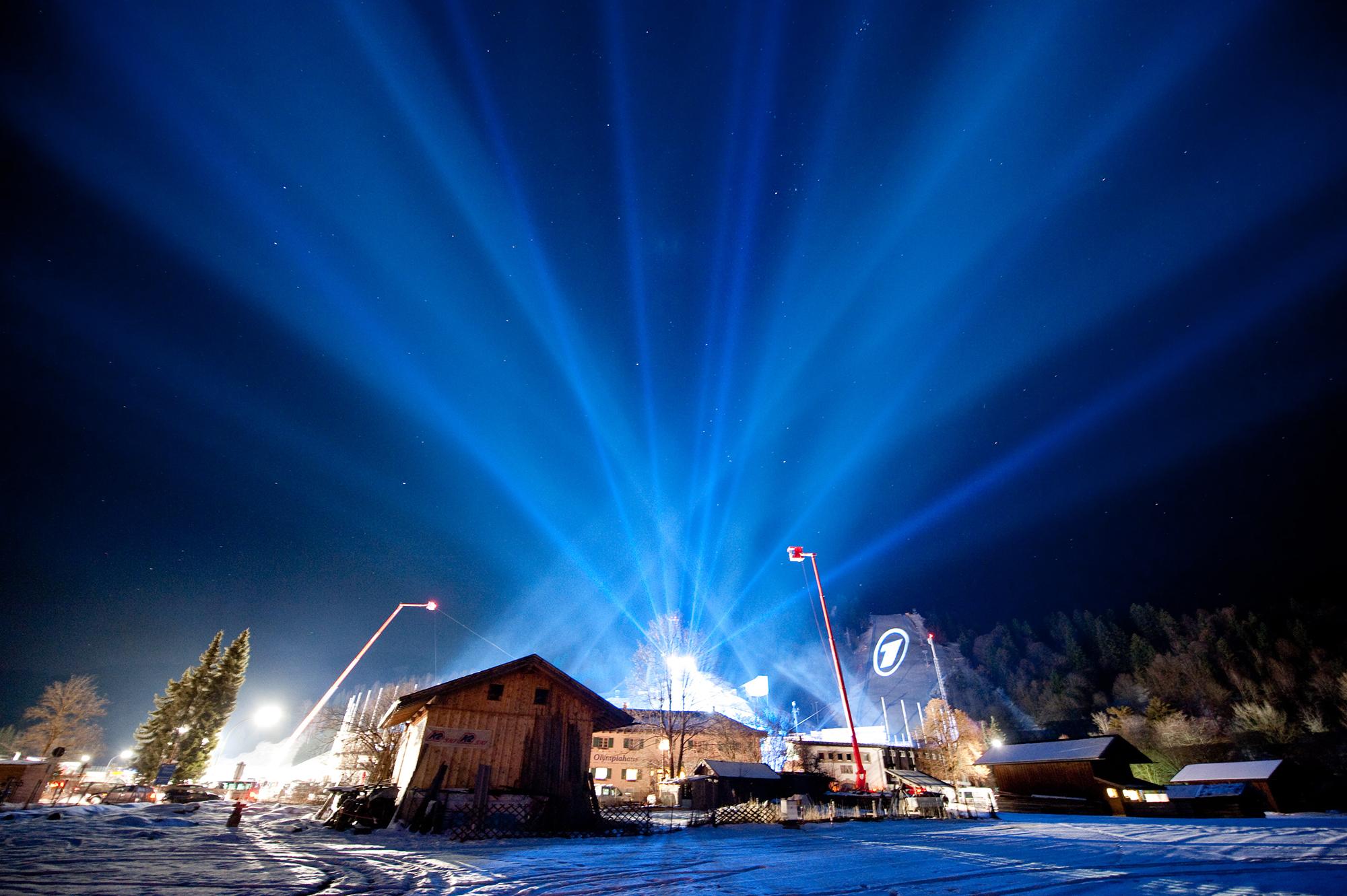 Skylights beam into the night sky. Snow is on the ground and we see a cluster of wooden buildings.