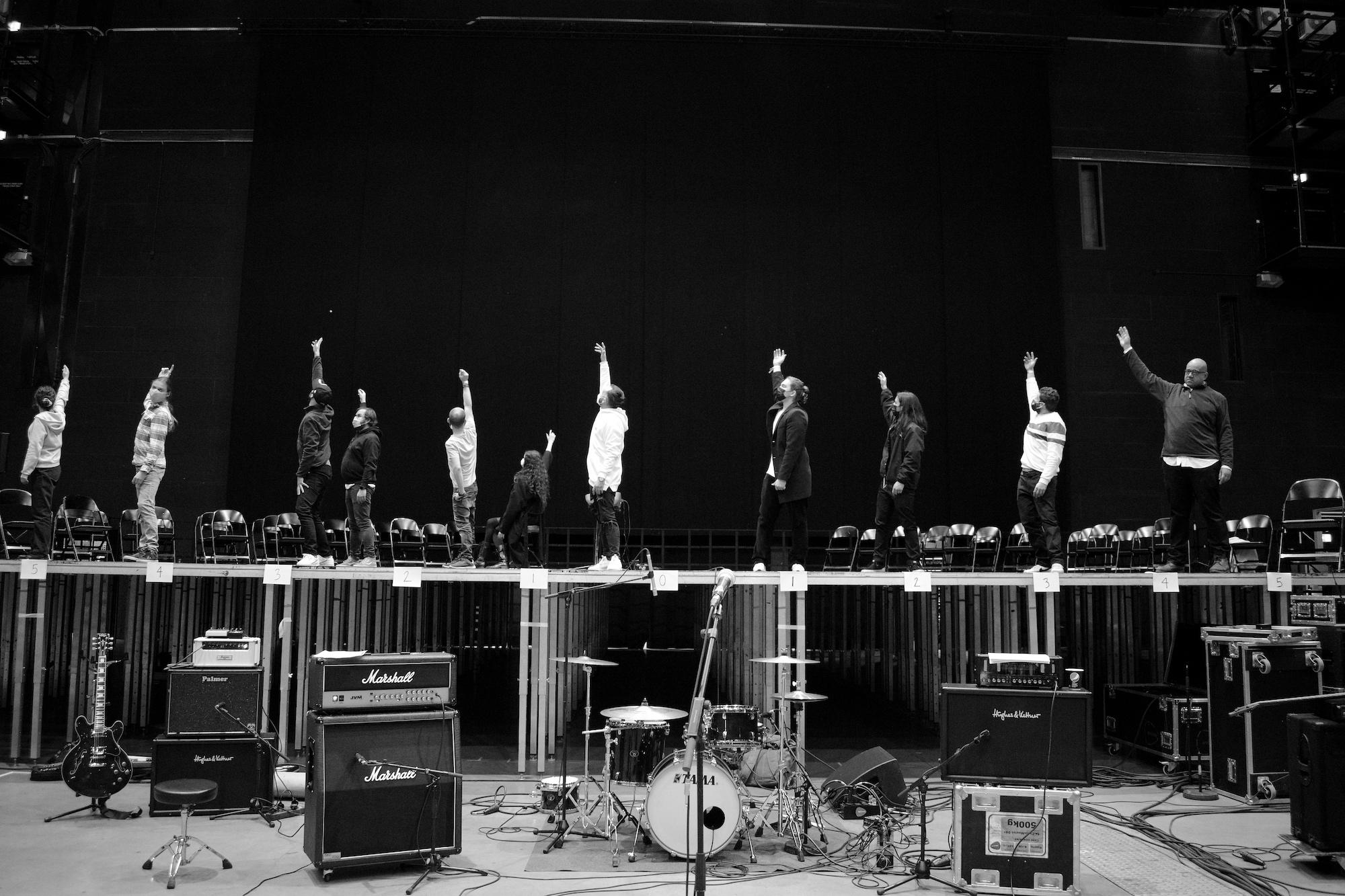 11 people stand on stage rehearsing for a performance. They all have their left hand in the air.