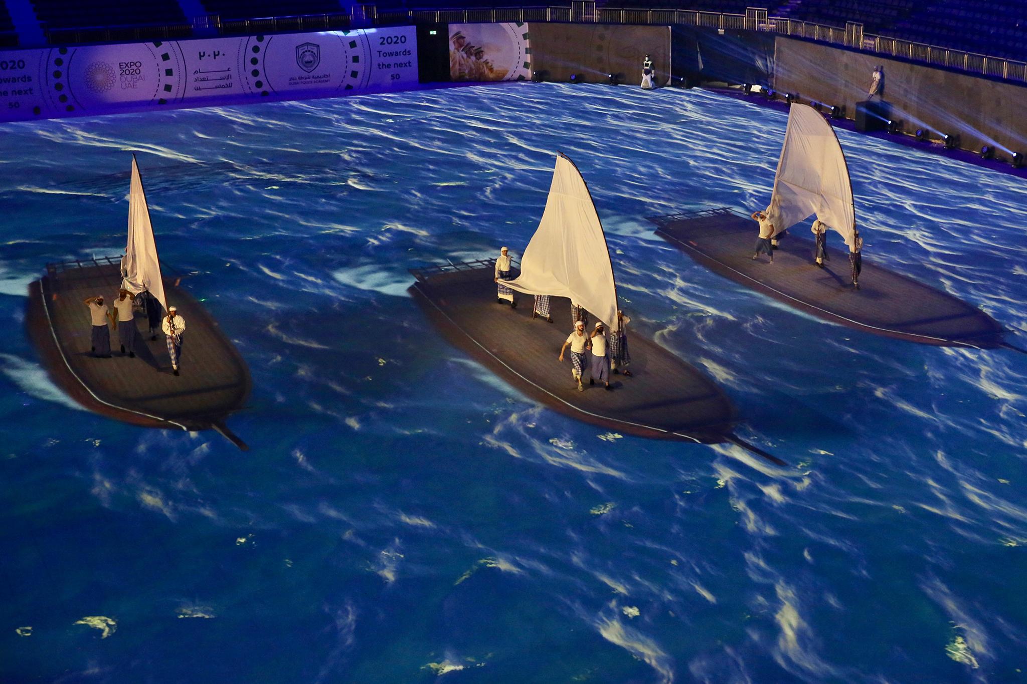 3 groups of men in white shirts hold physical cloth sails on top of video mapped and projected boats and ocean.