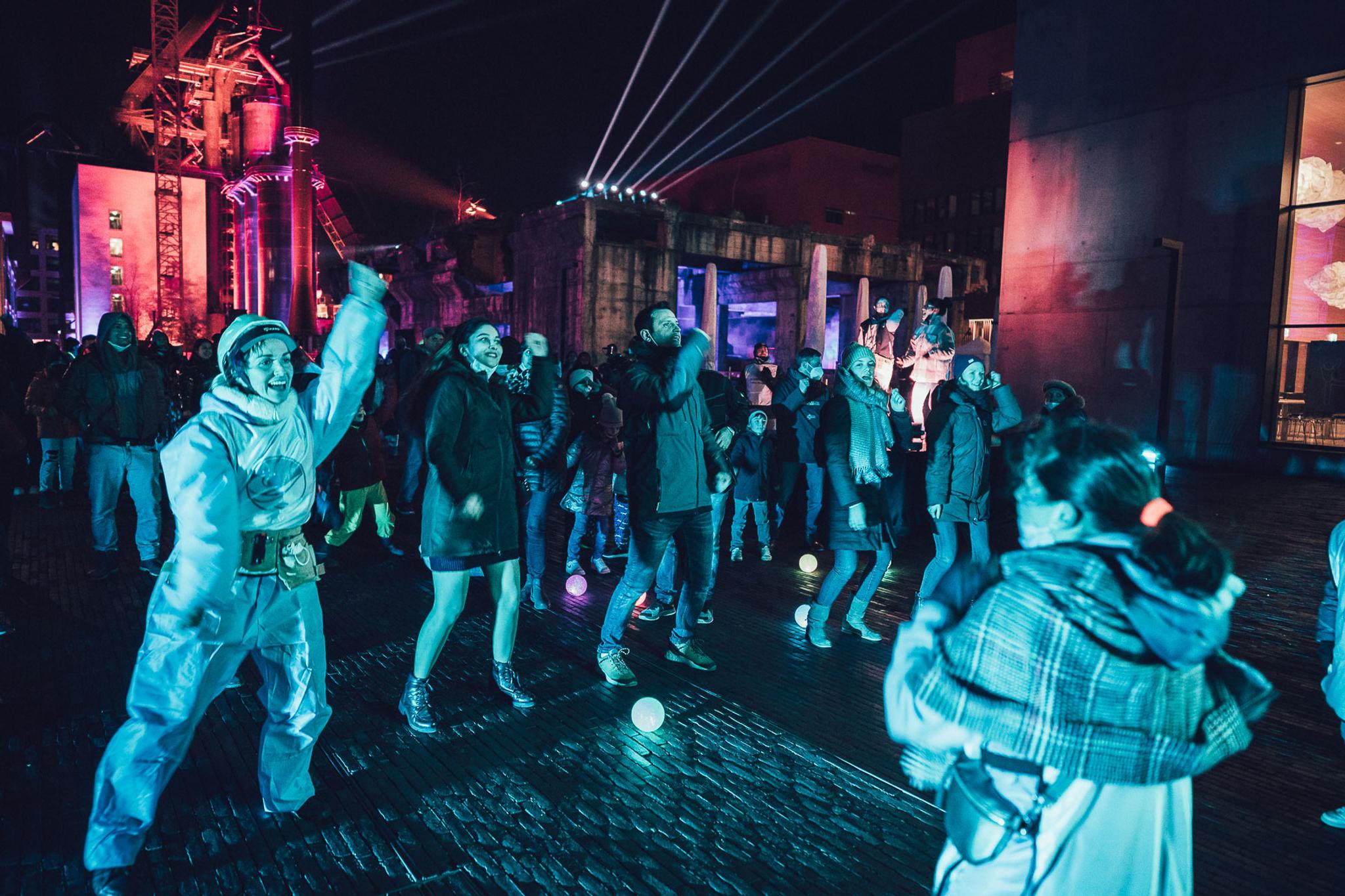 A group of people lit in blue light wearing winter coats imitate the movements of a woman in a puffy full body suit. They all raise their right arms up and we see they are in movement.