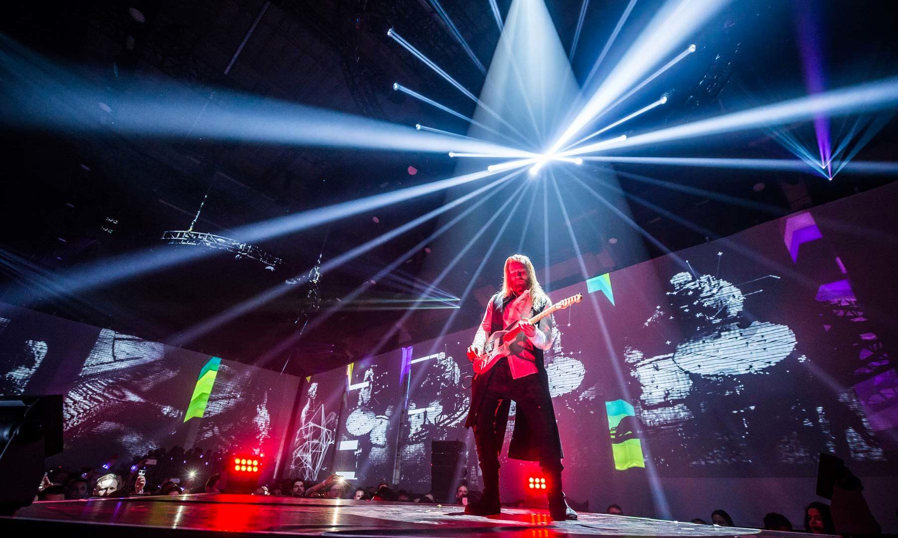 A guitarist plays on stage surrounded by video mapping and spotlights in a club environment.