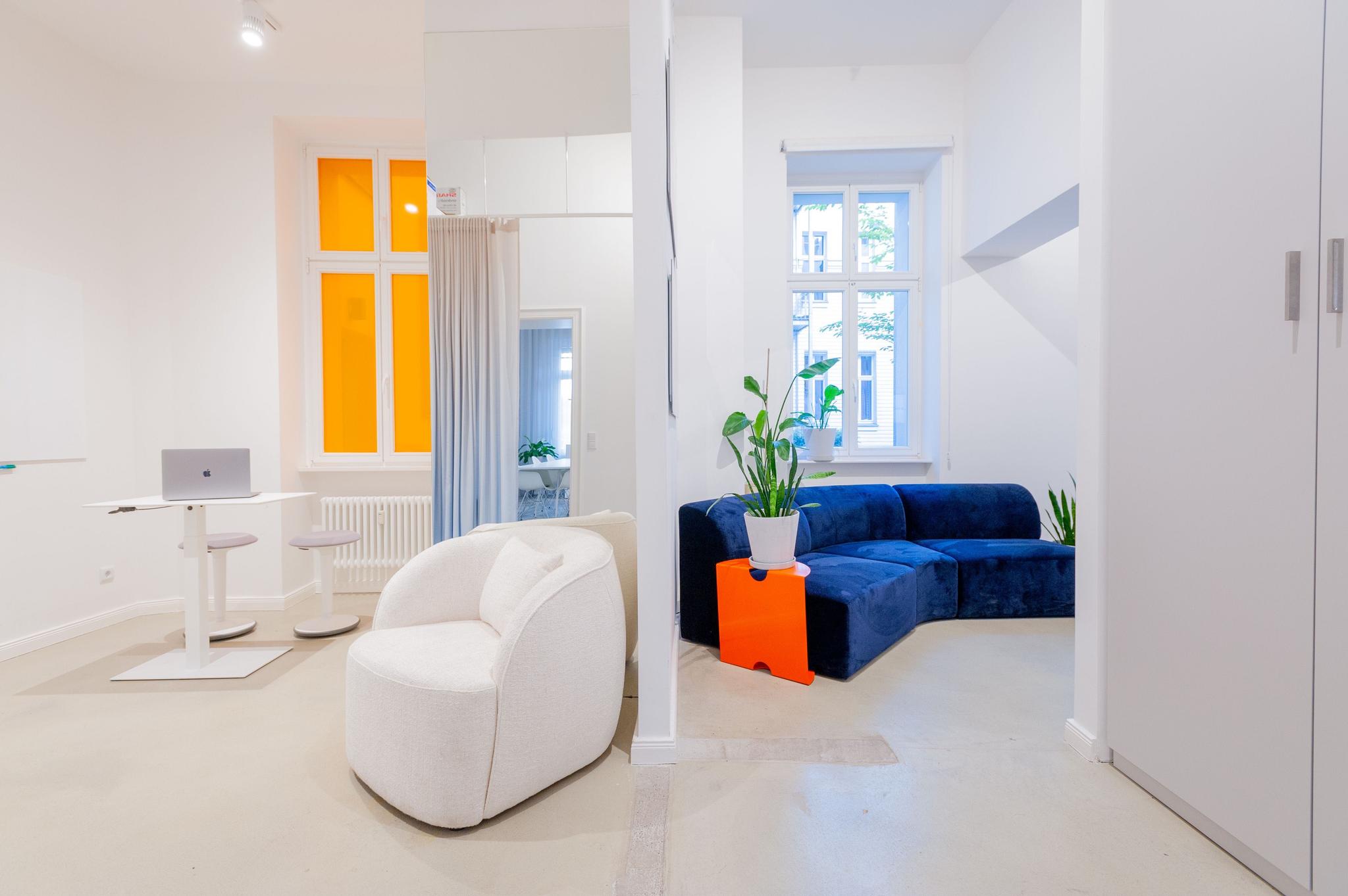 White, open office space with accents of orange and blue. To the left we see a standing desk with a macbook on it. On the right next to a wall-divider, there is a deep blue curved couch with a potted plant next to it.