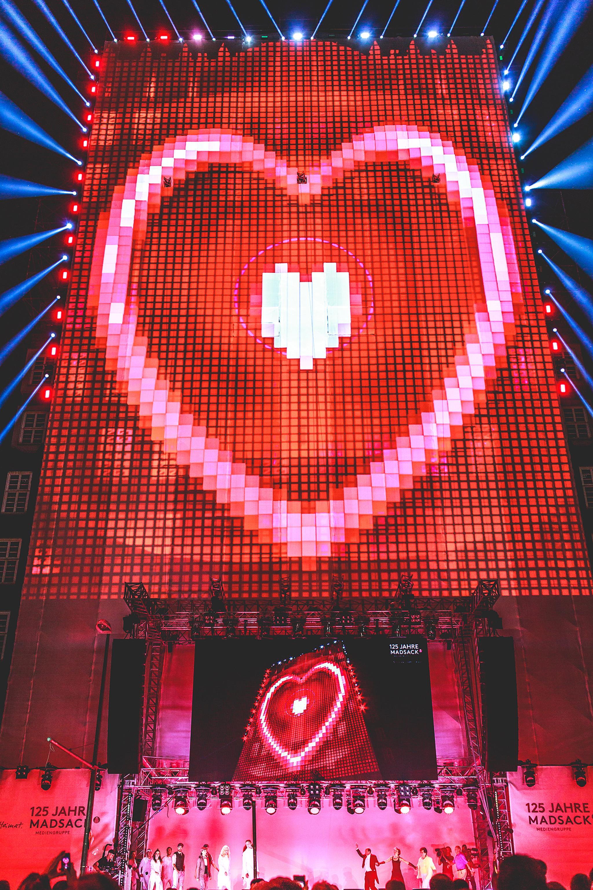 Video mapping of a bright, pink heart on a large vertical stage. A large crowd watches a musician outdoors.