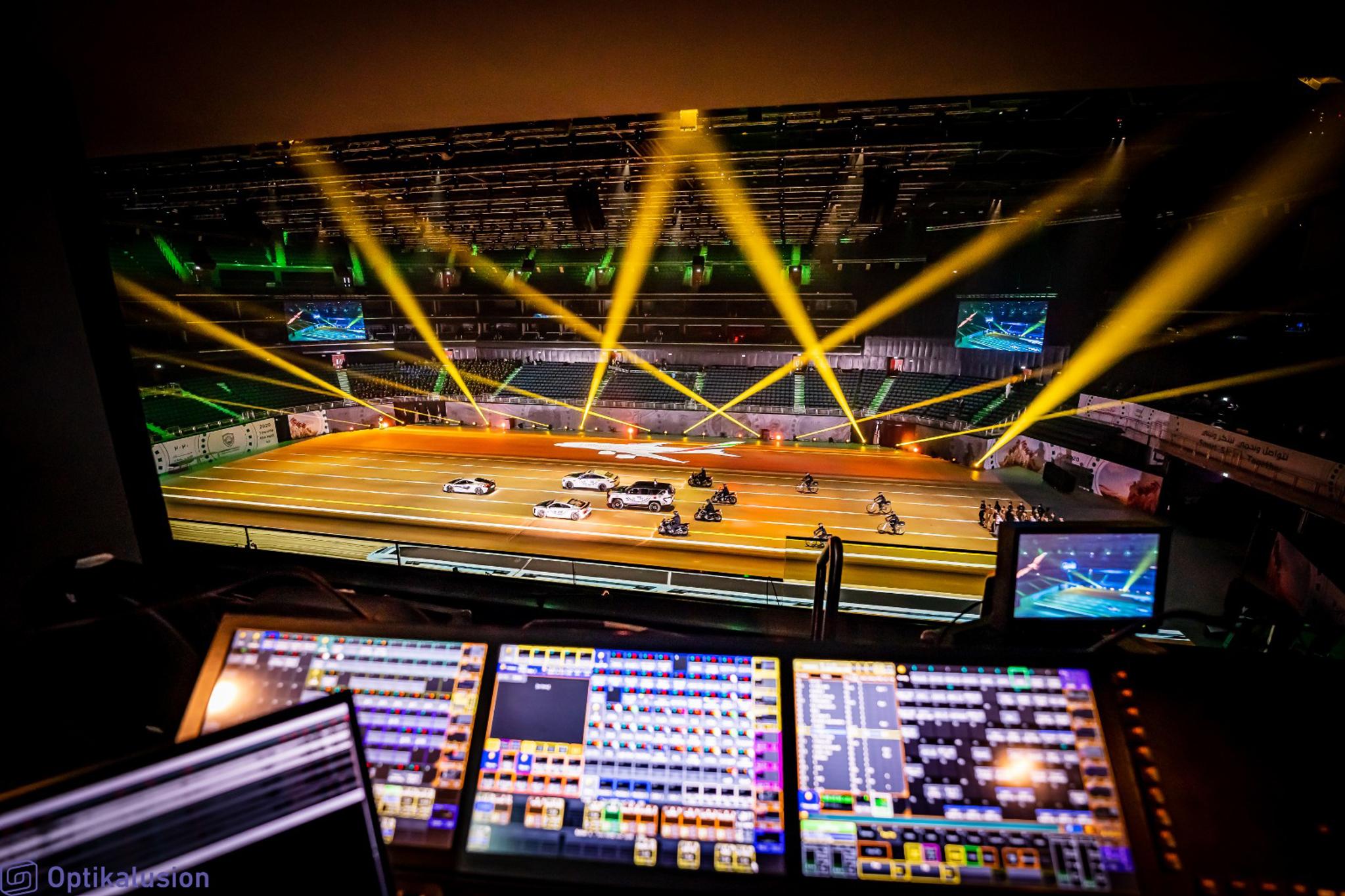An indoor arena as seen from the tech booth. Beams of orange spotlights emit from the ground and cars drive over projection mapped content on the ground.