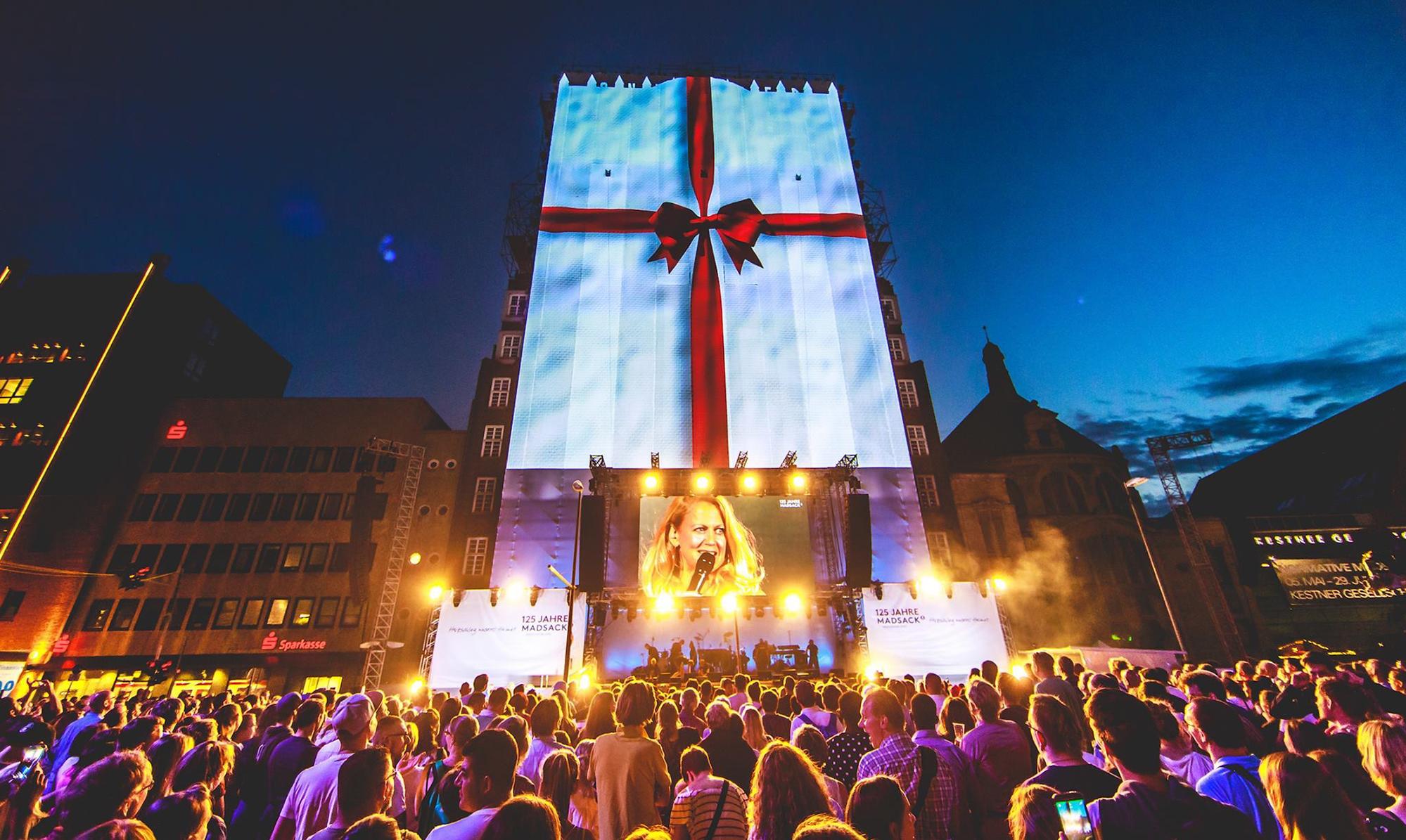 Video mapping of a gift- white background and a red ribbon- on a large vertical stage. A large crowd watches the musician outdoors.