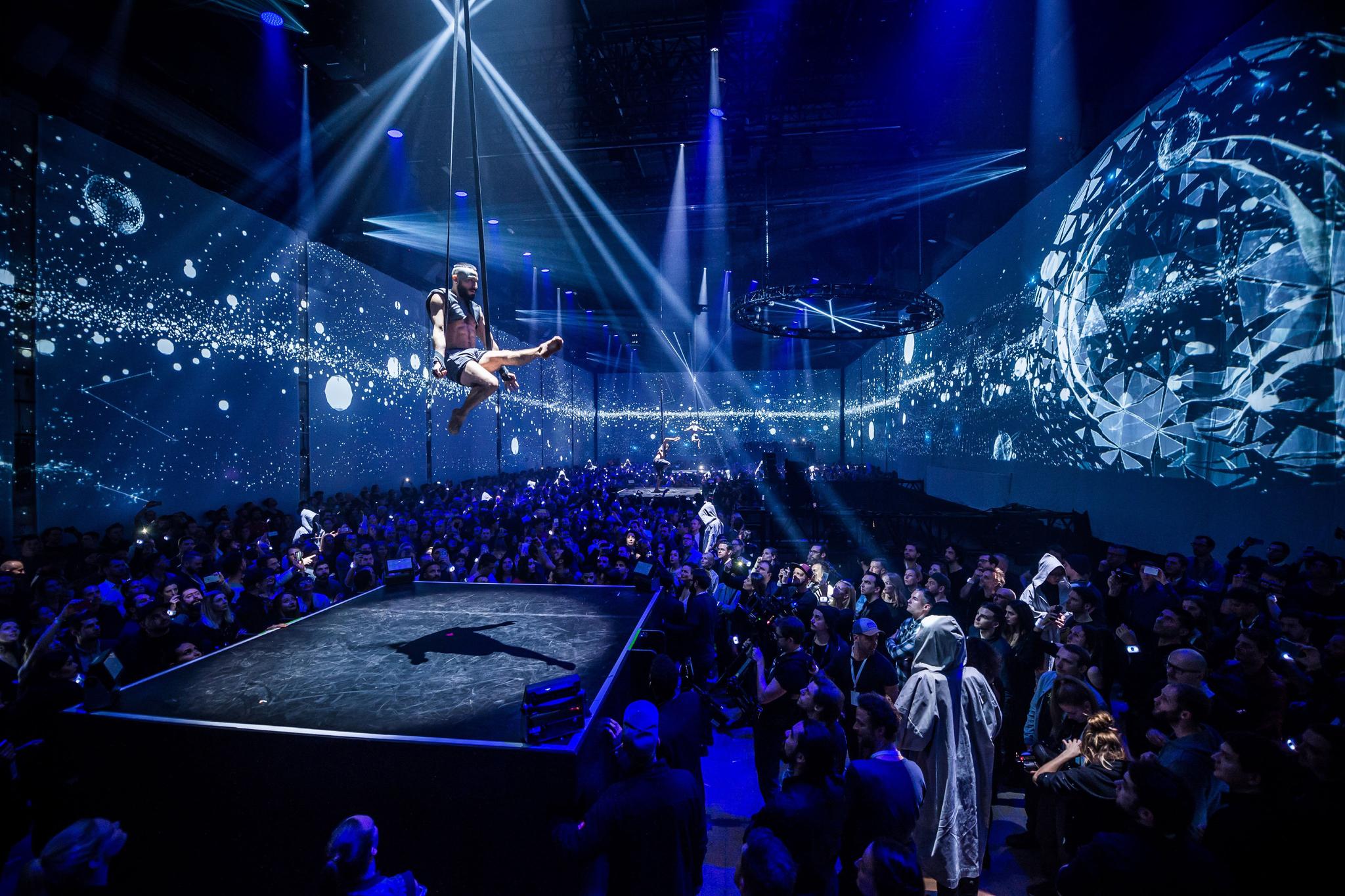 An acrobat performs flying above a crowd in a club setting. Projection mapping in blue on the walls.