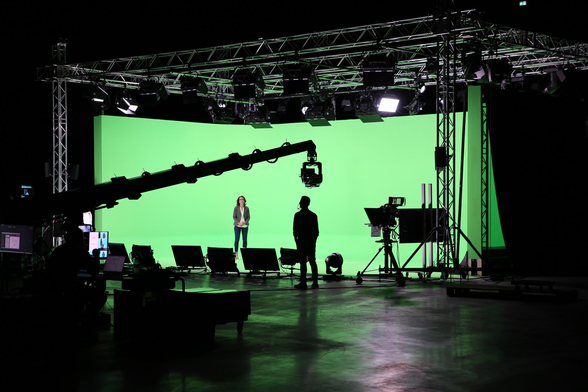 A long camera crane extends into a spacious, tall greenscreen which surrounds the subject who is wearing business clothes. The subject speaks into the camera and the director looks at them, wearing a headset. Many lights are on the ceiling, fixed to metal trusses.
