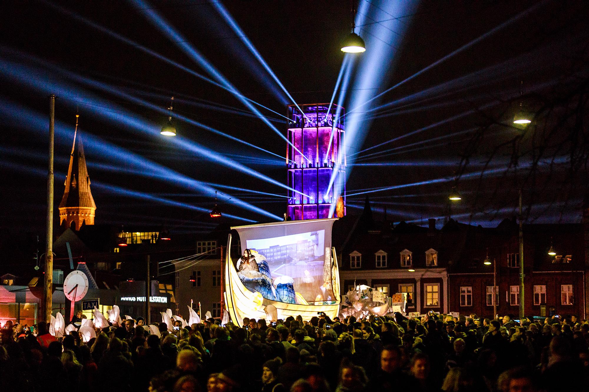 The lantern boat makes its way through the crowd as a purple and pink lit tower throws spotlights into the night sky.